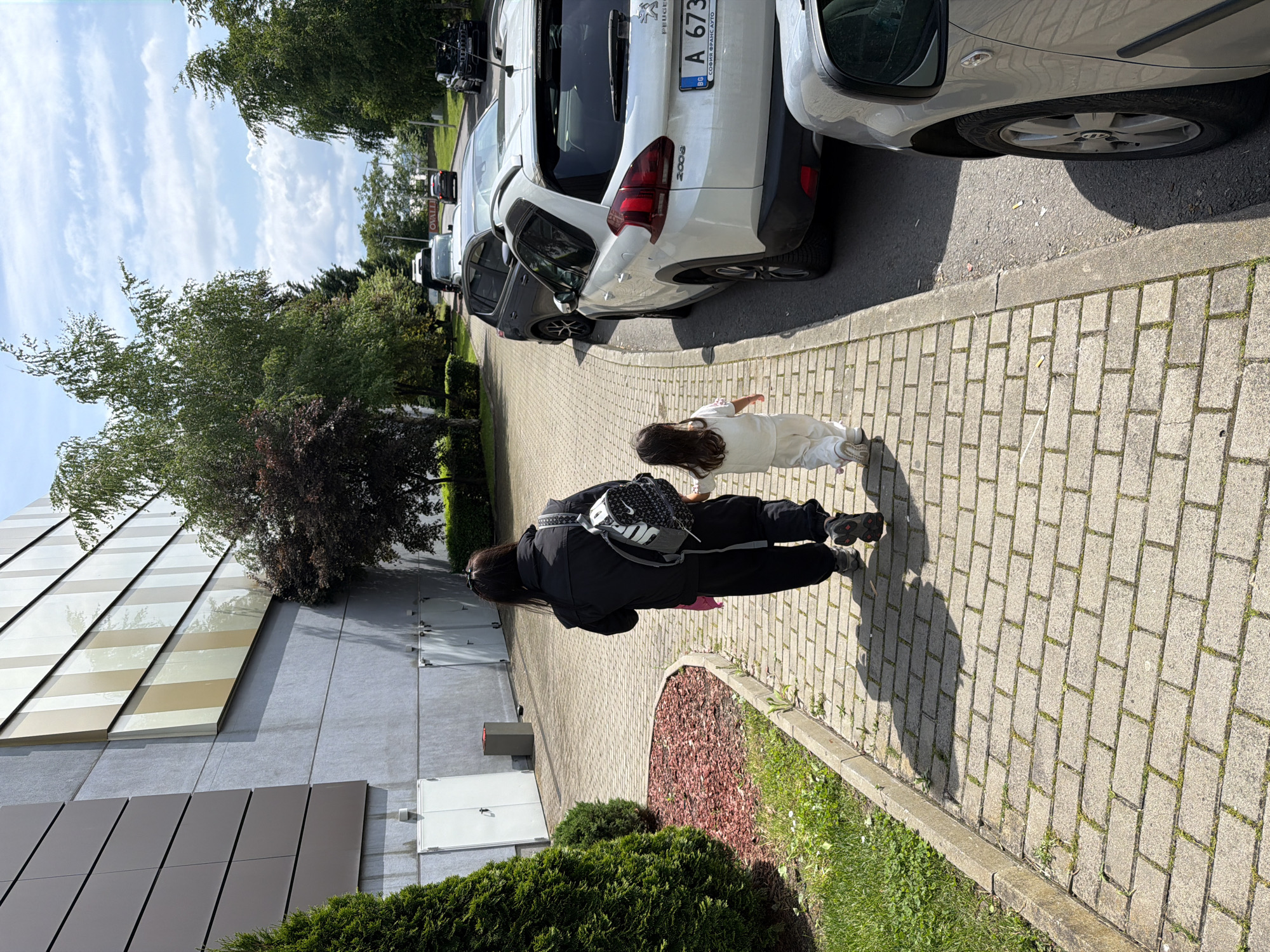 A child and an adult walk on a paved sidewalk in Burgas, Bulgaria. The child, wearing white clothing, extends her arms as if flying. The adult, clad in black clothing and carrying a backpack, walks beside her. Cars line the adjacent street, including a white Peugeot with Bulgarian license plate number A 6733. The street and sidewalk are flanked by trees and bushes. A building with patterned windows is also visible.
