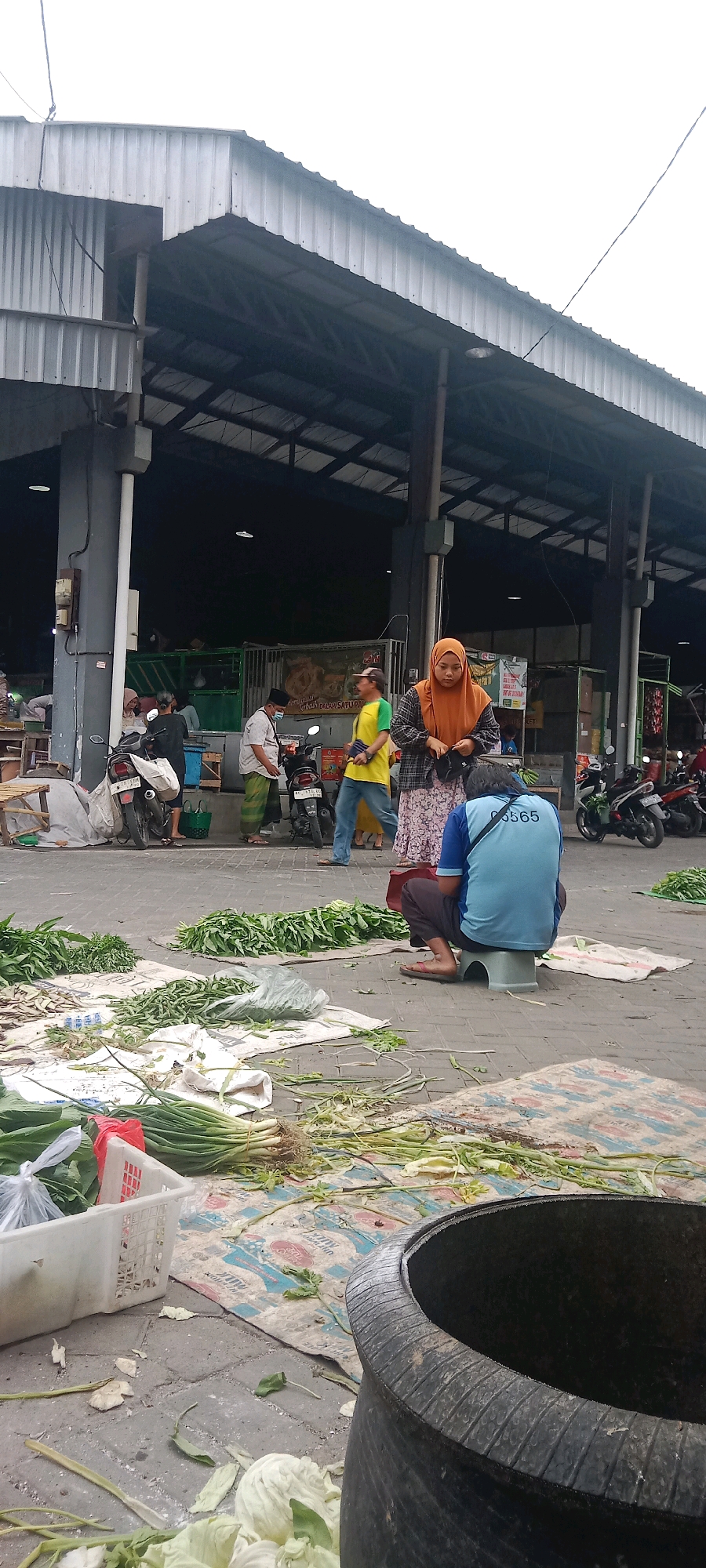 The image depicts an outdoor market scene in Kras, Indonesia. A woman in an orange hijab is standing and adjusting her clothing, facing a person who is sitting on a small stool, facing away from the camera. This seated individual is wearing a blue t-shirt with "0565" printed on the back, and dark pants. In the foreground, various vegetables, including green leafy produce and spring onions, are spread out on cloths on the ground. Several motorcycles are parked in the background. Behind the motorcycles, there's a covered market structure with a metal roof and supporting pillars. Other individuals are visible in the middle ground, some appearing to be vendors or shoppers. The environment suggests a typical bustling local market setting.