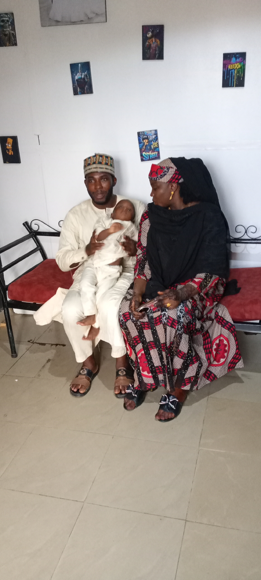 Three people, a man, a woman, and an infant, are seated on a bench indoors. The man, dressed in a light-colored robe and cap, is holding the infant, who is clad in a similar light-colored garment. To his right, the woman is seated, wearing a patterned dress with a black shawl and headwrap. She is holding a smartphone. The bench has a dark metal frame with a red cushion. The background features a white wall adorned with various framed images. The location appears to be in an interior space in the city of Unknown, Unknown.