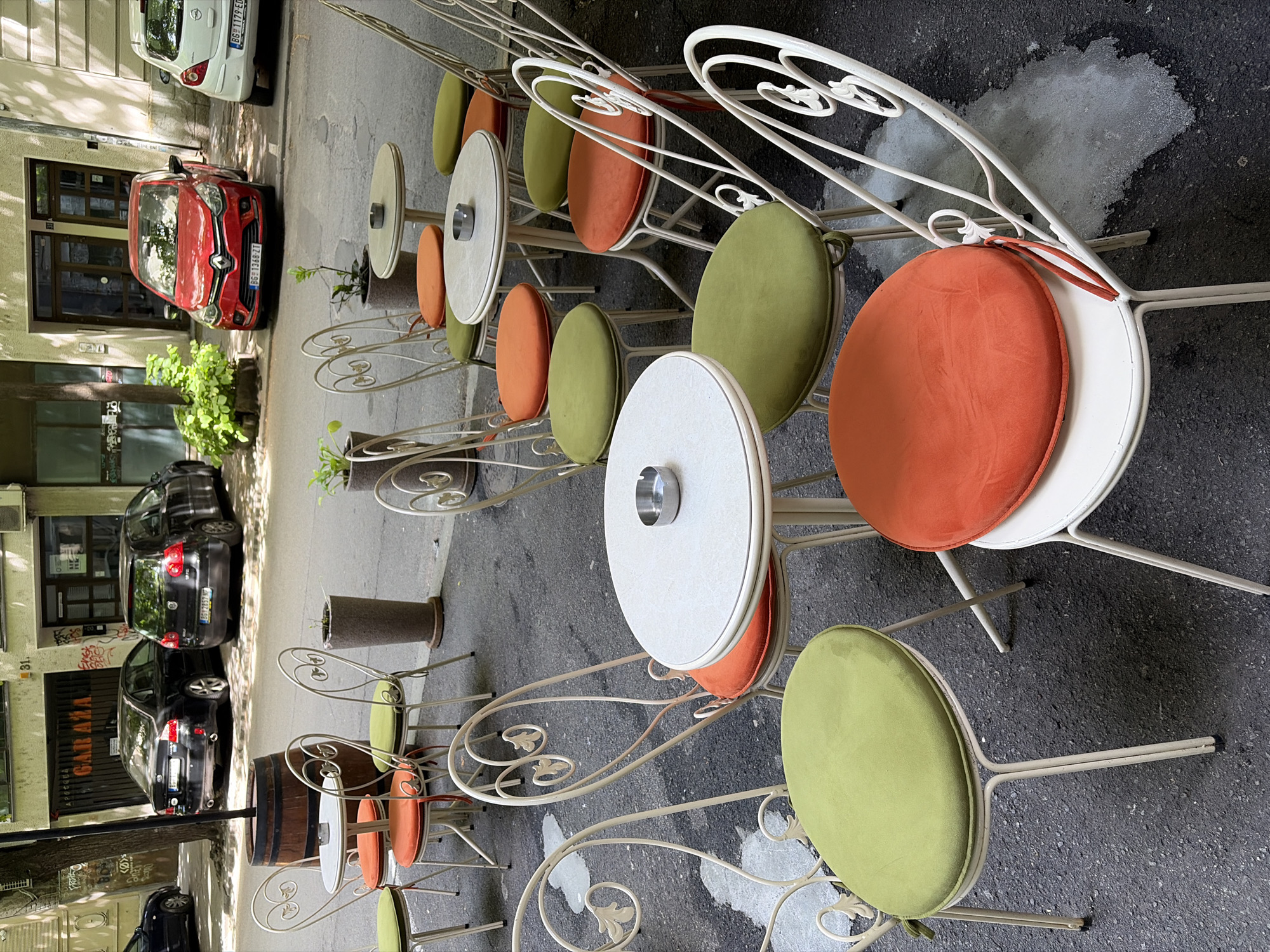 Outdoor cafe setting in Belgrade, Serbia. Several rows of metal tables and chairs, alternating orange and green cushions. Tables feature a central ashtray. Cars parked along the side of the street, buildings visible in the background.
