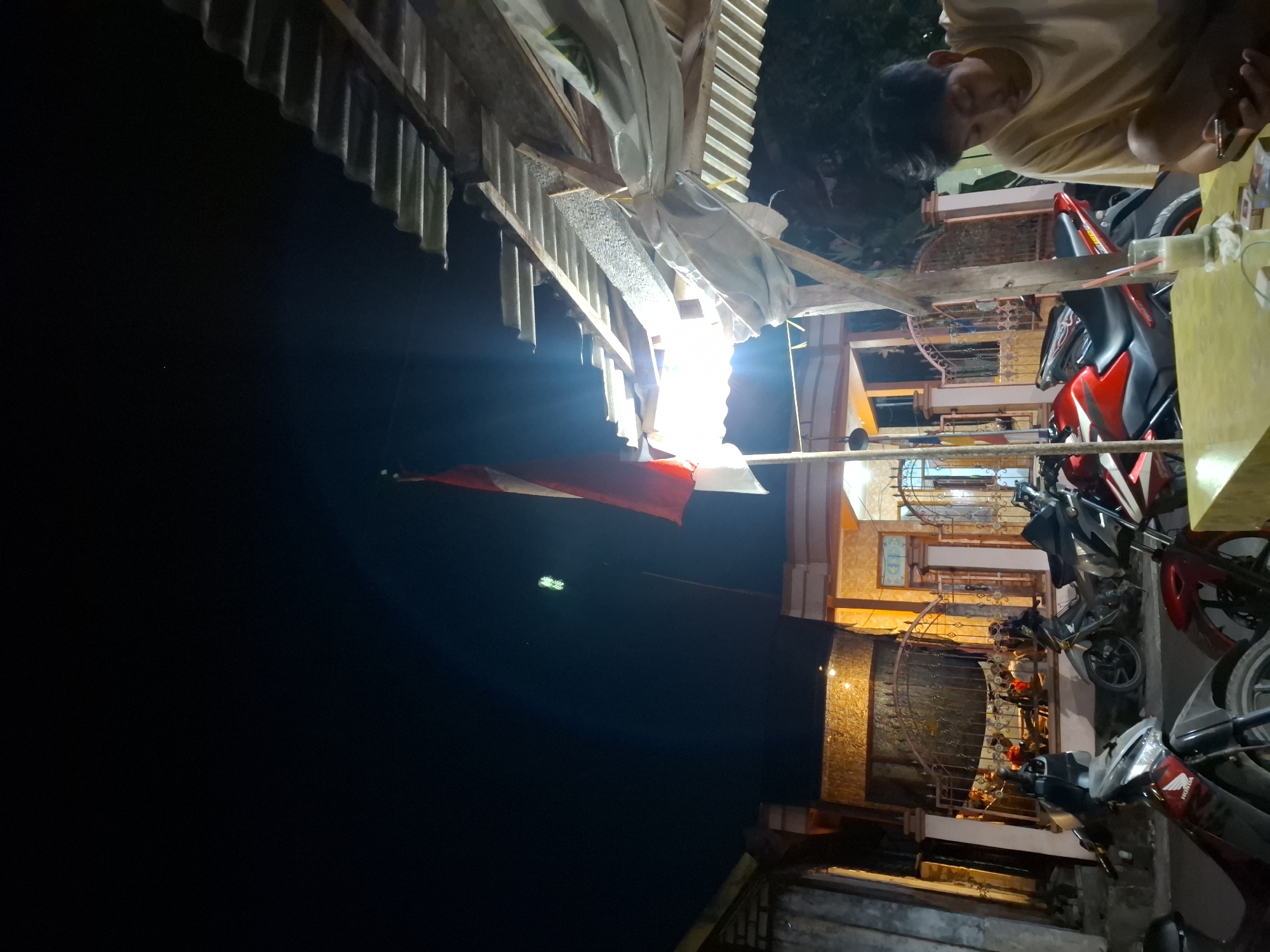 In the city of Bulubrangsi, Indonesia, at night, an Indonesian flag with red and white stripes is flown from a pole, partially obscured by a corrugated metal roof structure supported by wooden beams. To the right, a young man wearing a light-colored t-shirt sits at a yellow table, looking down at a phone in his hand, with a pack of cigarettes and a glass containing a light-colored beverage nearby. In the background, several motorcycles are parked in front of a building with illuminated windows and a decorative metal fence. The scene is dimly lit, with artificial lights illuminating the area.