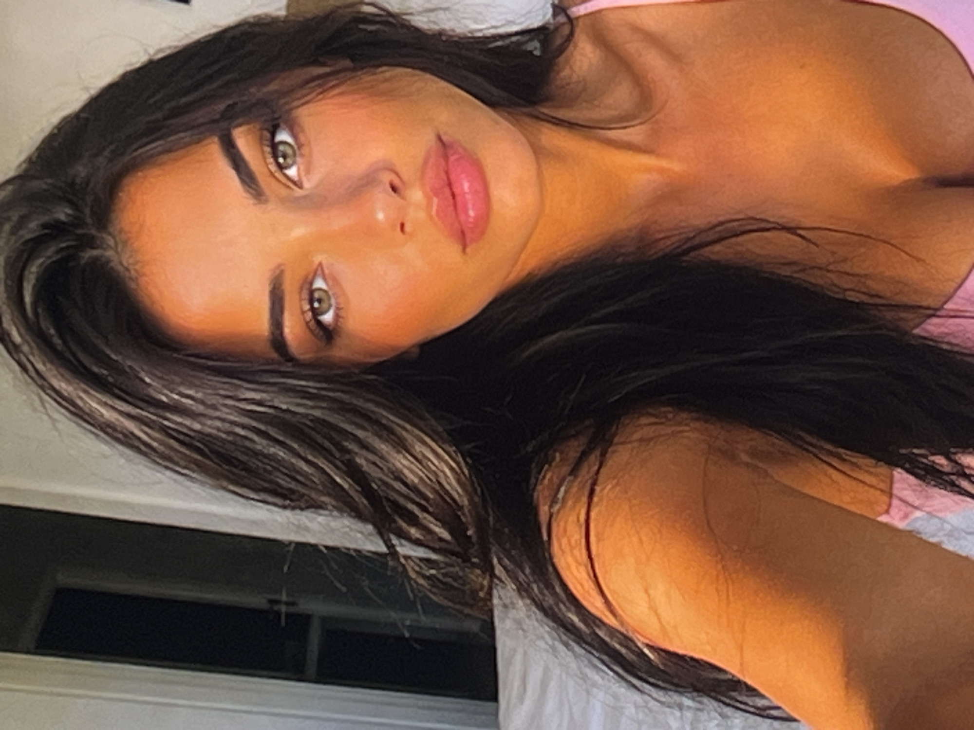 A close-up, selfie-style shot shows a young woman with dark, wavy hair framing her face. Her eyes are a striking green, and her lips are full and pink. She appears to be wearing a pink top, with the neckline visible. The lighting in the image is warm and golden, possibly indicating late afternoon sun. The background is softly blurred, with hints of a white surface, possibly a bed, and a doorway with a dark interior. The overall impression is one of natural beauty and relaxed intimacy.