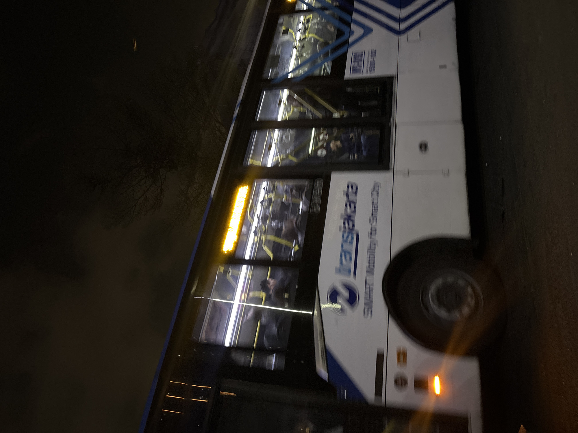 A side view of a white Transjakarta bus is captured at night in North Jakarta, Indonesia, as it moves past the camera. The bus features a prominent blue and white logo and lettering that reads "Transjakarta" and "SMART Mobility for Smart City." The windows of the bus are illuminated from within, revealing the blurry silhouettes of passengers seated inside. Yellow poles and handles are visible through the glass, typical of public transportation interiors. The bus's headlights cast a warm glow, and an orange indicator light is visible on the lower front portion of the bus near the wheel. The background is dark, with the suggestion of trees and a hazy sky, indicating an outdoor urban environment at night. The overall impression is one of motion, with the slight blur of the bus and its surroundings suggesting it is in transit.