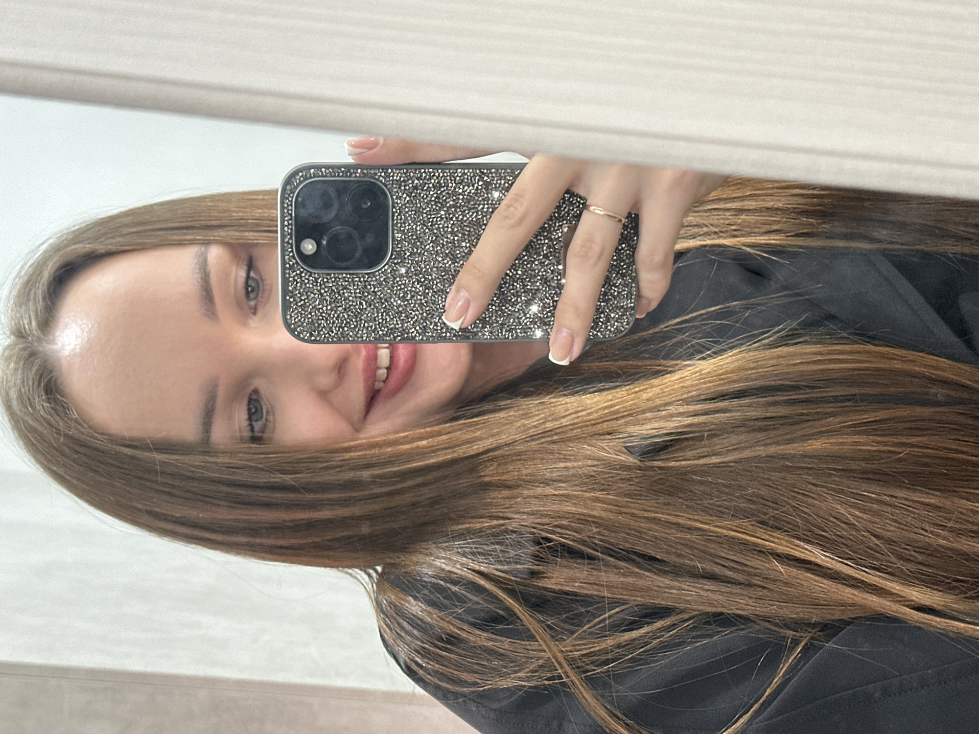 A young woman is taking a selfie in a mirror with her smartphone. The phone has a dark case covered in small, reflective studs or crystals. She's holding the phone in her right hand, which also features a thin, gold-colored ring. Her nails have a French manicure. She is smiling at the camera. She is wearing a dark-colored top. She has long, brown hair with highlights. The background is a plain white or light-colored wall, seemingly indoors. The location is not discernible from the image, other than it being in a room with light-colored walls and possibly a mirror. There is no indication of the city of Chaikovsky, Russia.