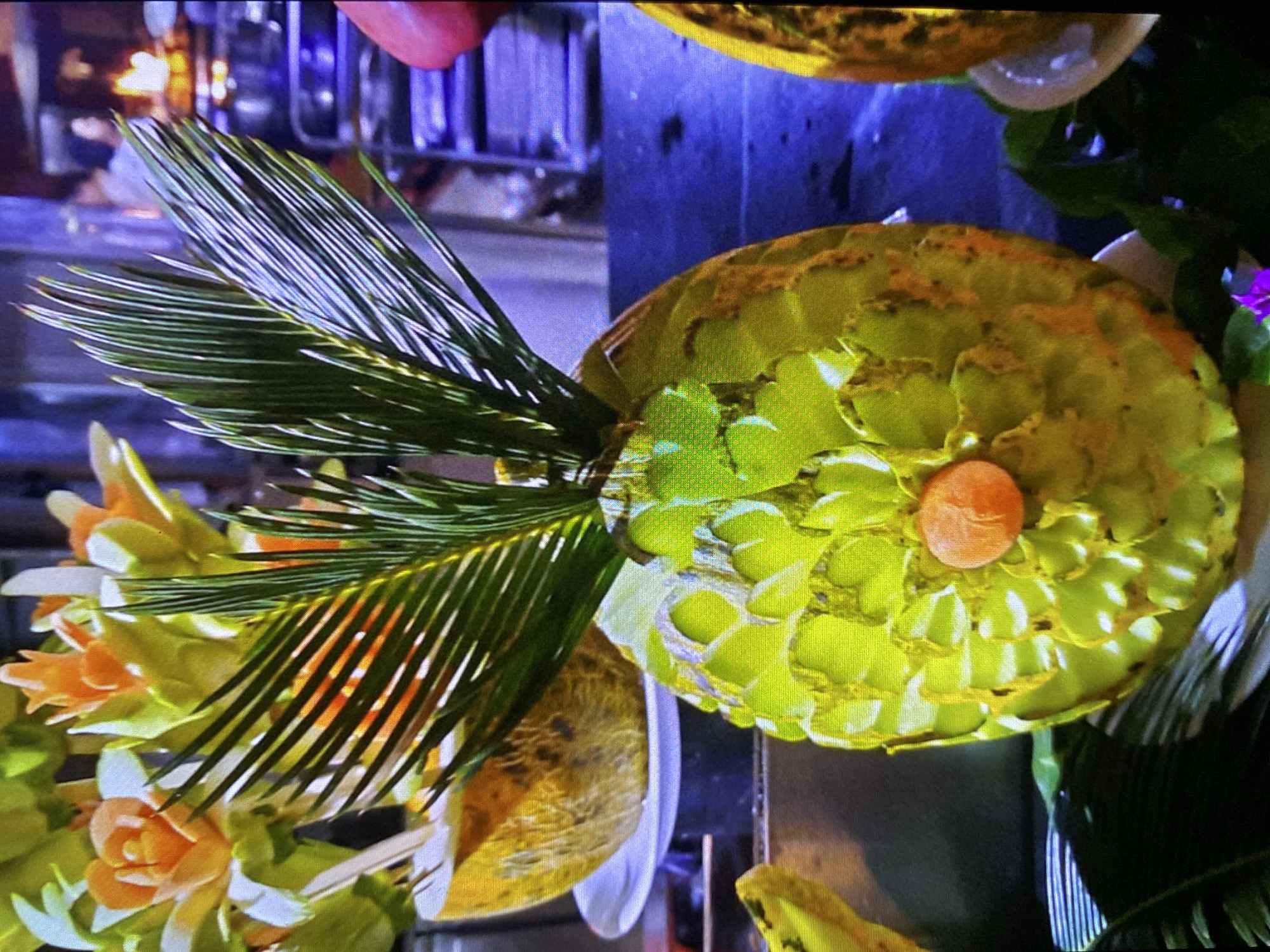 The image displays an elaborate fruit carving, likely a large melon such as a watermelon or honeydew, serving as a decorative centerpiece. The main carved fruit, oriented centrally and facing right in an upright view, features intricate patterns of light green and yellow, resembling layered leaves or scales, with a small, round orange piece at its core.
To the left of this central piece, a dark green palm frond extends dynamically, adding a natural element to the arrangement. Below and slightly to the left, a cluster of smaller, delicately carved floral designs in shades of orange, yellow, and white are visible, suggesting more fruit or vegetable artistry. Other carved fruits, less distinct, appear partially in the upper right and lower left corners, contributing to a larger display.
The setting appears to be an indoor venue, possibly a buffet, banquet, or event space, with blurred metallic and blue-tinted elements in the background hinting at professional kitchen equipment or display stands. The lighting is artificial, illuminating the vibrant colors of the carved items. The image captures a moment of food art presentation, showcasing meticulous craftsmanship. There is no visible text. The context indicates the location is Bankya, Bulgaria.