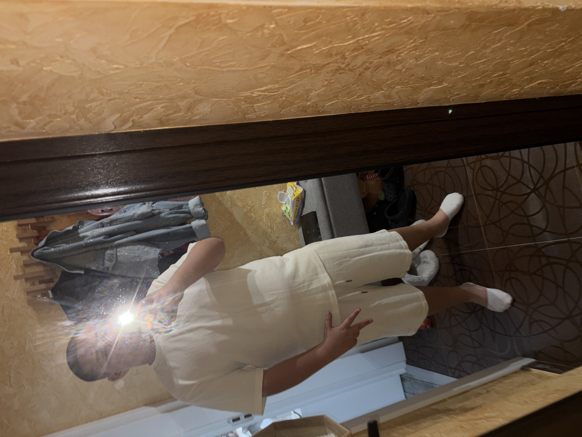 A male individual, dressed in white clothing and white socks, stands in an interior space that appears to be a room within a residence. The man is gesturing with his hand, displaying a peace sign. The room features patterned brown tile flooring. Other objects present in the room include clothing, shoes, and furniture, all situated against a backdrop of textured walls. The location is presumed to be in a private residence, potentially in Shumen, Bulgaria.