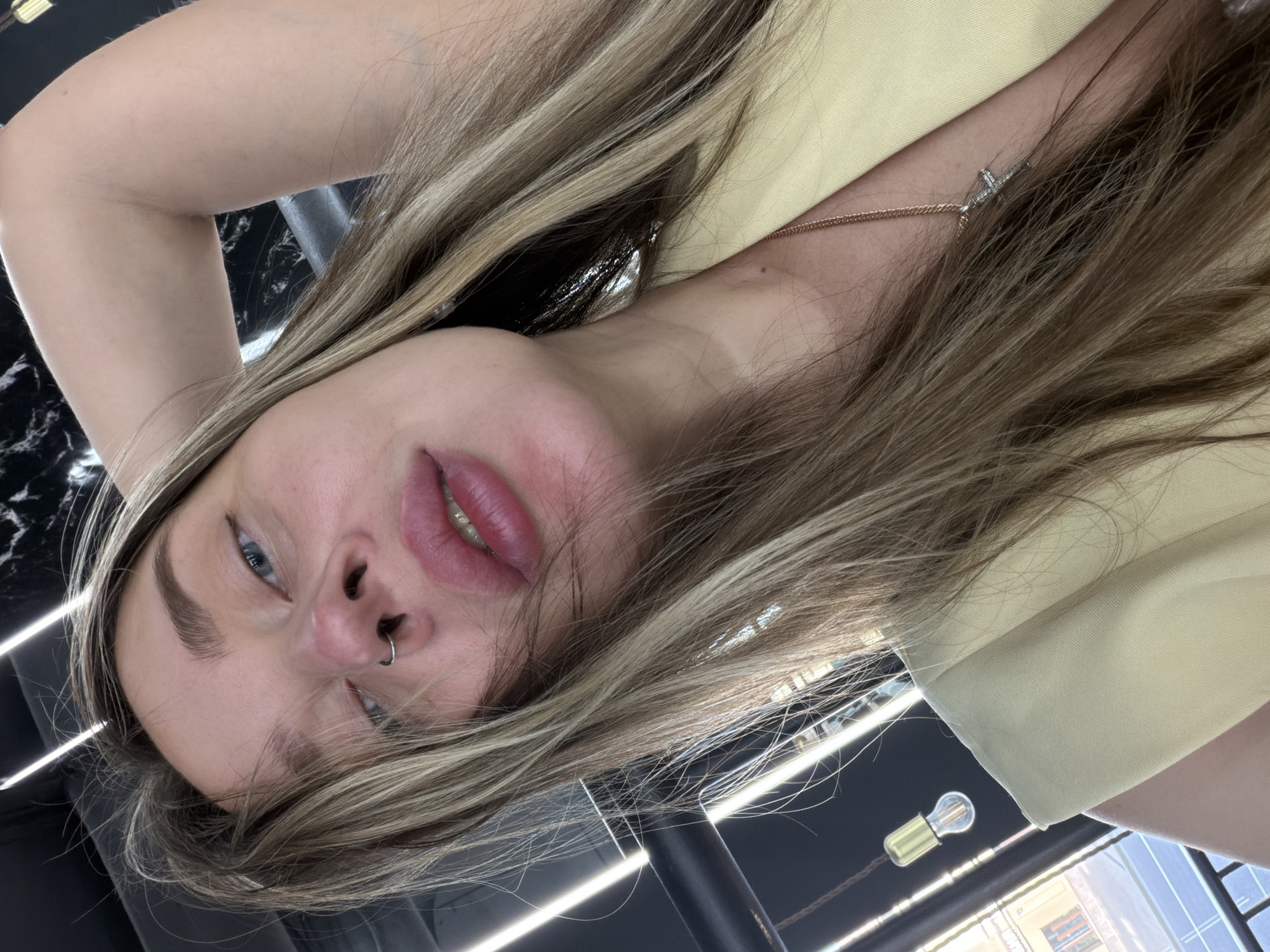A young woman with blonde hair and a nose piercing is photographed at an unusual angle. She has blue eyes and is wearing a light yellow top. A gold necklace with a pendant is visible. The location appears to be a beauty salon in Moscow, Russia, evidenced by the illuminated ceiling fixtures, salon equipment in the background, and glimpses of Moscow's urban landscape through a window.