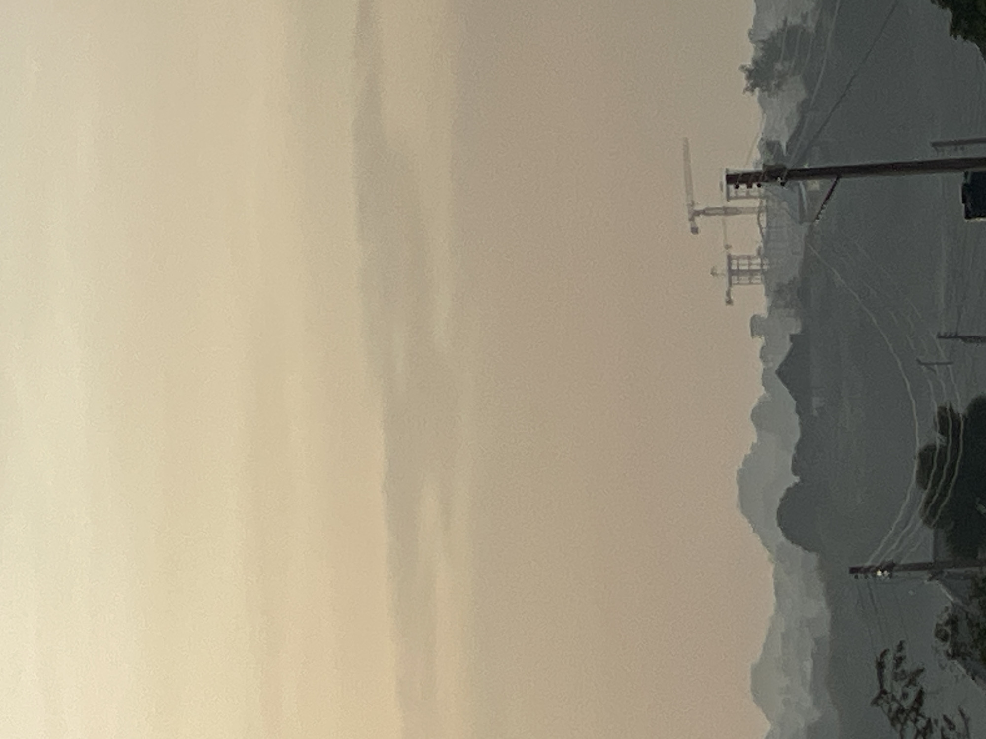 The image, when viewed upright, displays a wide, hazy landscape, likely in Jos, Nigeria, under an overcast sky or during a period of significant atmospheric haze (such as Harmattan dust). The upper portion of the frame is dominated by a pale, nearly uniform light beige or cream-colored sky, indicating diffused light and low visibility.
In the mid-ground, several large, dark structures resembling construction cranes or industrial equipment are silhouetted against the hazy backdrop. Further into the background, indistinct outlines of rolling mountains or hills are visible, their forms softened and muted by the atmospheric conditions.
On the right side of the foreground, several utility poles with multiple power lines extending horizontally across the frame are prominent. Some dark green foliage from trees or bushes can be seen along the very bottom and right edge.
No people or discernible text are present. The time of day appears to be either early morning, late afternoon, or an overcast midday, characterized by soft, diffused light and a lack of clear shadows. The overall scene conveys a sense of industry within a natural, expansive, and somewhat obscured environment.