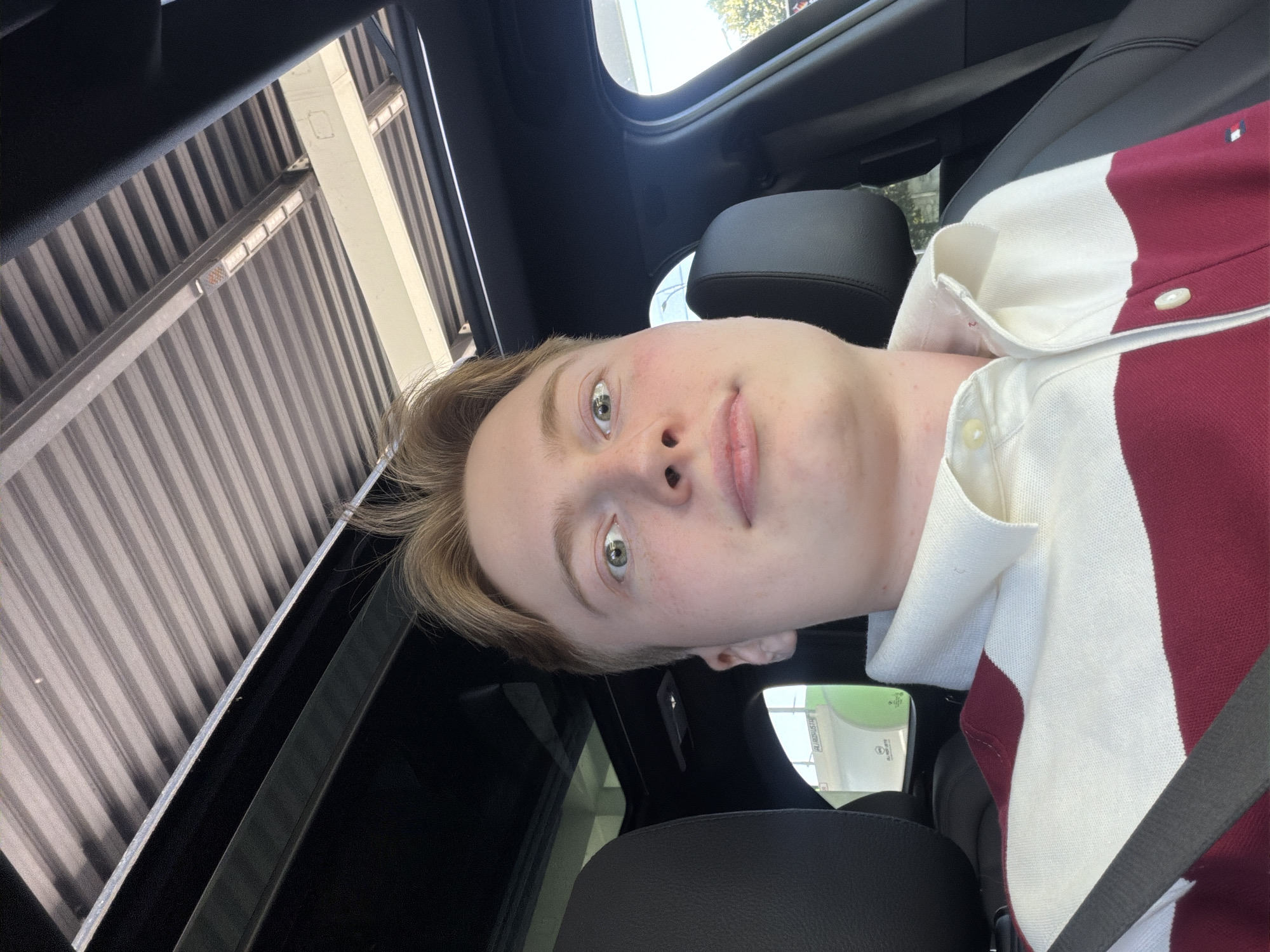 A close-up, selfie-style, over-the-shoulder shot shows a young white male with blond hair and light skin sitting in the passenger seat of a car. He is wearing a collared shirt with horizontal red and white stripes. The car's sunroof is open, revealing a ribbed ceiling structure above. In the background, through the car window, a green sign with text is visible, suggesting an outdoor location, possibly a commercial area. The angle of the shot indicates the photographer is holding the phone at a high level, pointing it downwards and towards themselves. No specific landmarks in Kyiv, Ukraine are identifiable from the provided image.