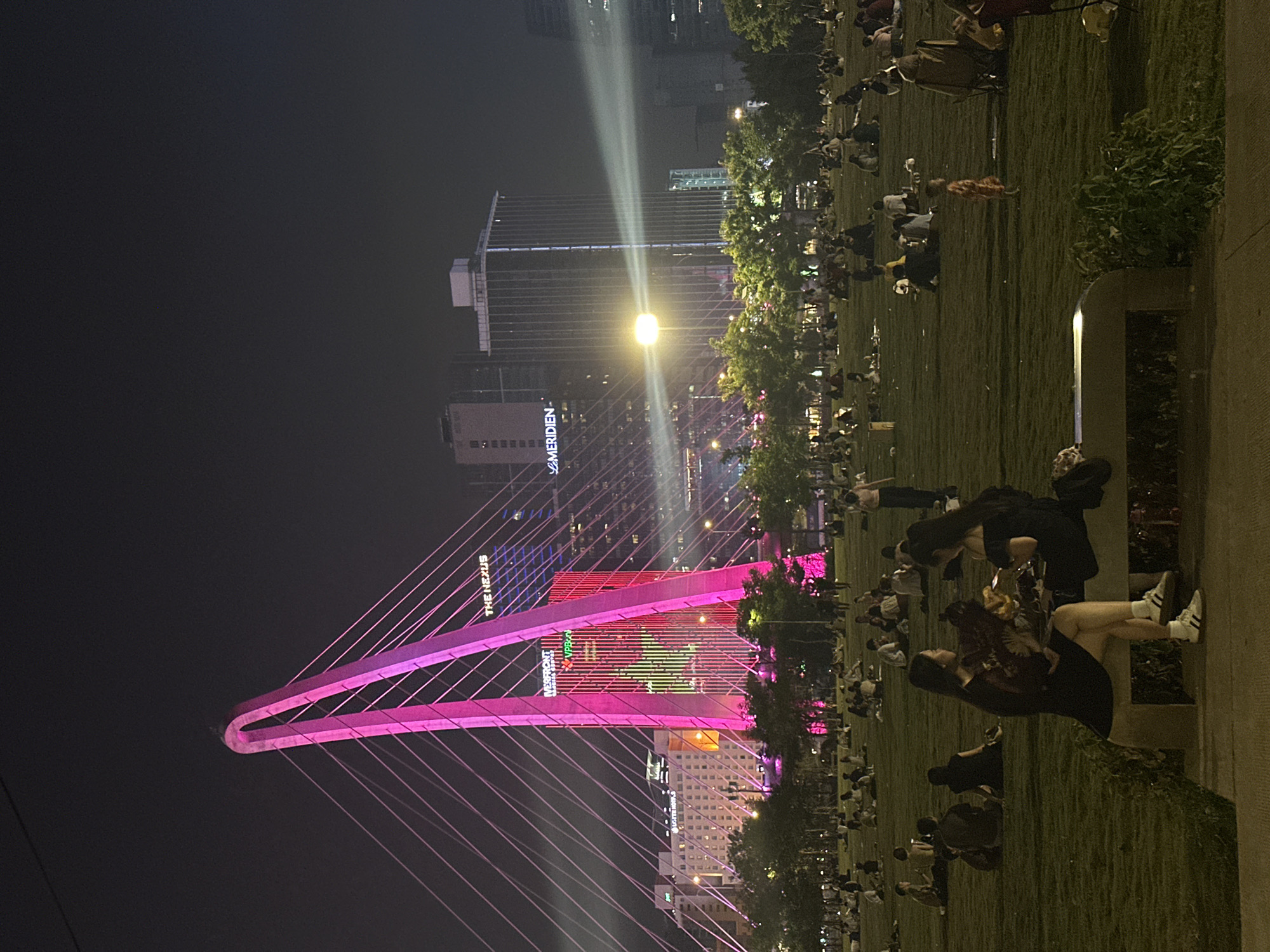 The image captures a vibrant night scene in an urban setting, likely a park or waterfront promenade. The primary subjects are numerous people gathered on a large grassy area, some sitting on the ground, others on what appear to be low benches or ledges. In the foreground, two women are prominently featured, one seated with her back to the viewer, the other standing behind her, both looking towards the illuminated cityscape.
The central activity involves people relaxing and observing the illuminated structures. The atmosphere appears casual and social, suggesting a gathering to enjoy the evening lights.
Notable details include a large, distinctive cable-stayed bridge, brightly lit in pink, forming an arch with numerous cables extending downwards. Behind the bridge, several tall buildings are visible. One prominent building has the text "MERIDIEN" vertically displayed, while another shows "THE NEXUS" and "VPBank" on its facade. Artificial lighting is a significant feature, with a powerful streetlamp emitting a strong beam of light, and buildings displaying various colored lights, including red, green, and blue patterns. Spotlights can be seen shining upwards into the night sky from the city. The time of day is clearly nighttime, with the darkness contrasting sharply with the bright artificial lights.