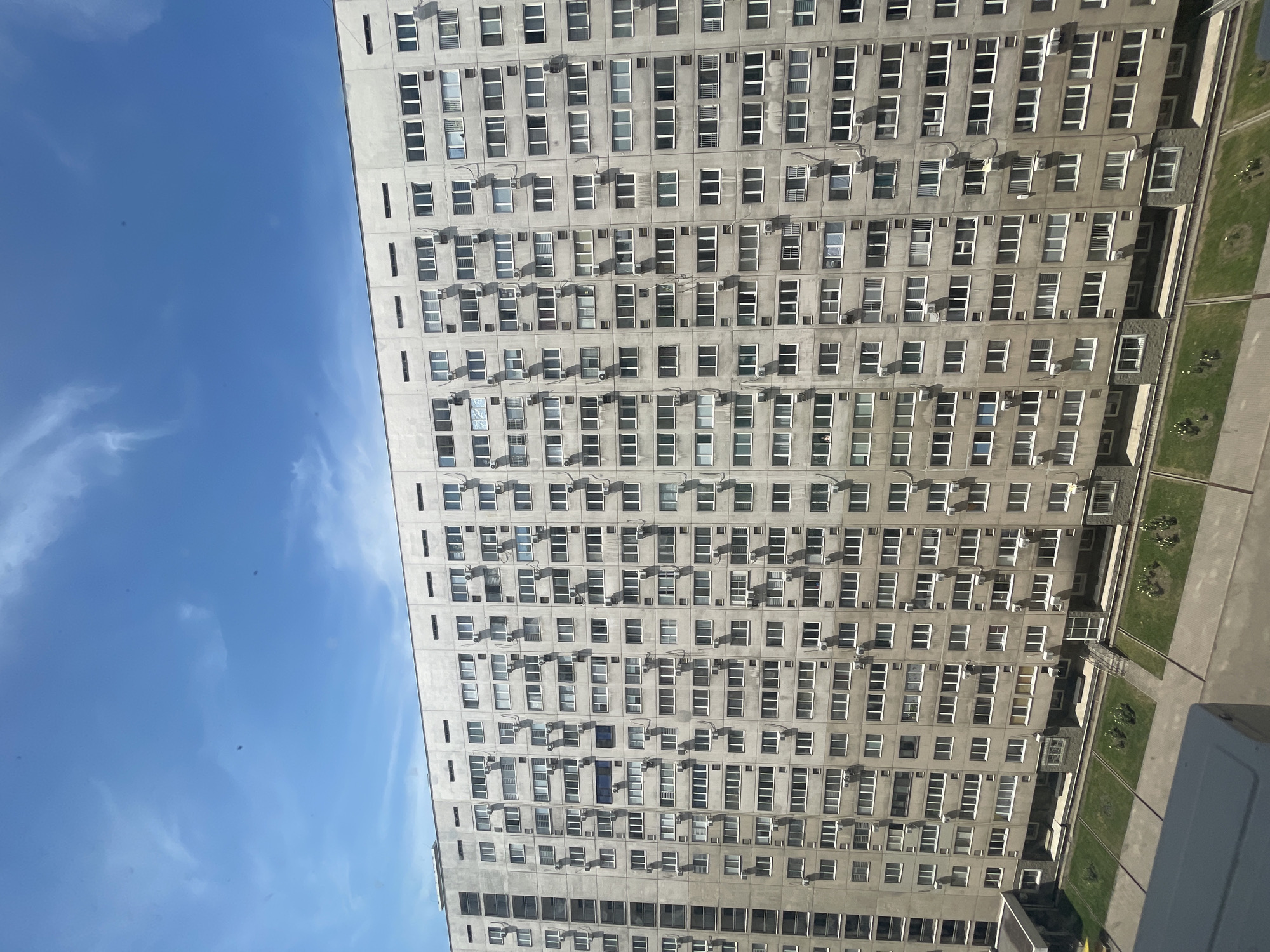 The image shows a large, multi-story residential building with a concrete facade, characterized by rows upon rows of identical windows and external air conditioning units. The building is viewed from a slightly elevated angle, revealing a section of its vast expanse. Below the building, there is a manicured lawn area with some small bushes and paved walkways. The sky above is a clear blue with scattered white clouds. The architectural style suggests it could be a Soviet-era mass housing block, common in many post-Soviet cities. Without specific identifying markers, pinpointing the exact location within Almaty is not possible based solely on the visual information provided.