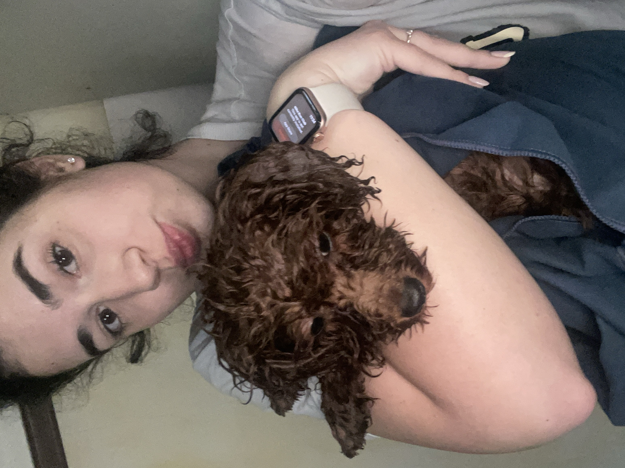 A person with dark curly hair and wearing an Apple watch is holding a wet, brown poodle dog in their arms. The person's left arm cradles the dog, which is predominantly in focus. Another dog is partially visible under a dark blue blanket. The background is a plain, light-colored wall. The location is not discernable from the image; there is no information to suggest it is in Sofia, Bulgaria.