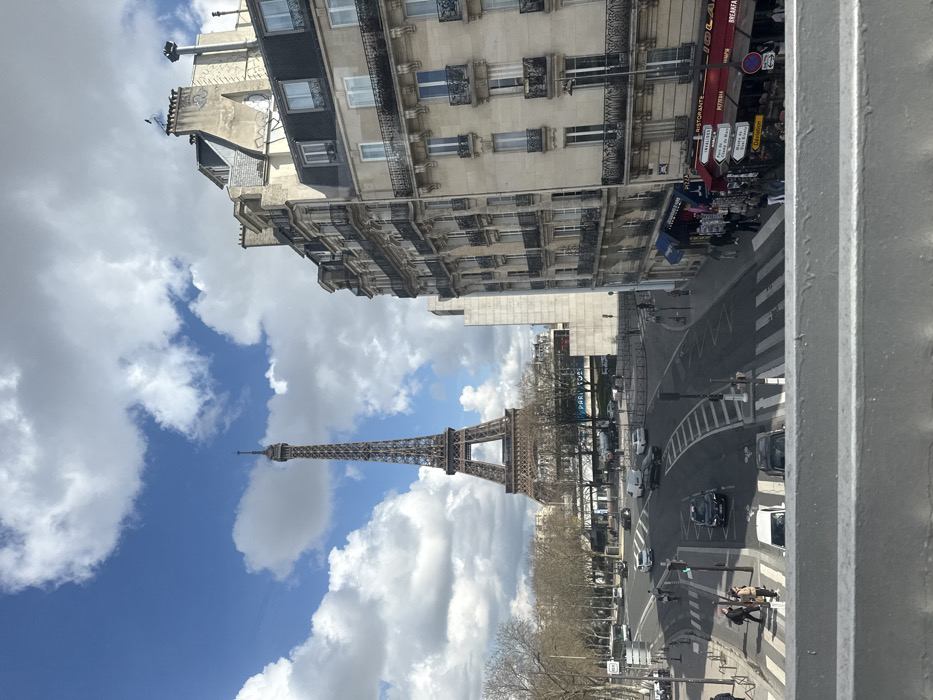 An outdoor wide shot captures a vibrant Parisian street scene on a partly cloudy day, featuring the Eiffel Tower prominently in the background. The vantage point is elevated, looking down over a multi-lane road intersection, with a gray railing visible in the immediate foreground.
In the center-left, the wrought-iron structure of the Eiffel Tower rises under a bright blue sky dotted with numerous white cumulus clouds. To its left, a park-like area with bare deciduous trees lines the far side of a wide boulevard, indicative of late winter or early spring. Several vehicles, including cars, a white van, and a white bus, navigate the curving asphalt roads, which feature clear white lane markings and pedestrian crosswalks. A motorcycle is visible to the left. Traffic appears moderate.
On the right side of the image, a multi-story, light-colored stone building, characteristic of Haussmann architecture, dominates the midground. It features numerous windows with intricate iron balconies and a dark gray mansard roof with dormer windows. The ground floor houses commercial establishments, including a shop with a blue awning selling goods and a "RISTORANTE" with a red awning and outdoor seating area. Pedestrians are scattered on the sidewalks and crossing the streets below, some congregating near the shops. A street sign cluster on the right points towards "INVALIDES" and "CHAMP DE MARS," aiding in location identification.
The scene depicts a busy intersection along a major thoroughfare in Paris, France, likely in the vicinity of the Quai Branly, looking towards the Eiffel Tower and the Champ de Mars area, consistent with a view from an elevated structure such as a bridge (e.g., Pont d'Iéna) or a building overlooking the Seine.
