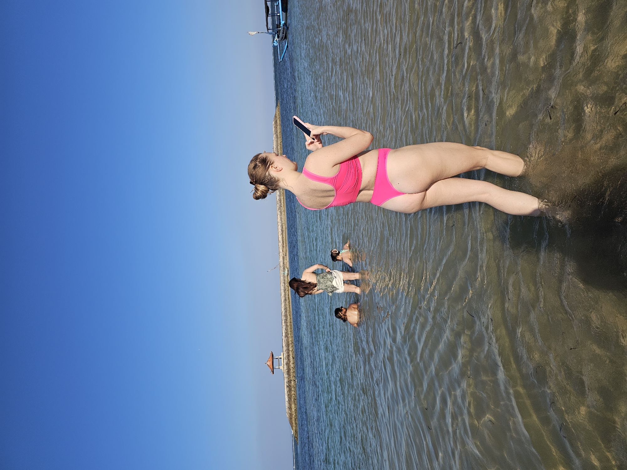 In Denpasar, Indonesia, on a clear day with a vibrant blue sky, four individuals are wading in the shallow waters of a beach. A woman in the foreground, facing away from the camera, is wearing a bright pink bikini and holding a smartphone. In the middle ground, three other individuals are also in the water. One person, facing away, wears a patterned top and white shorts, while two others are partially submerged. In the background, a stone breakwater with a small gazebo is visible, and a blue and white boat is moored offshore. The water is clear, revealing the sandy bottom.