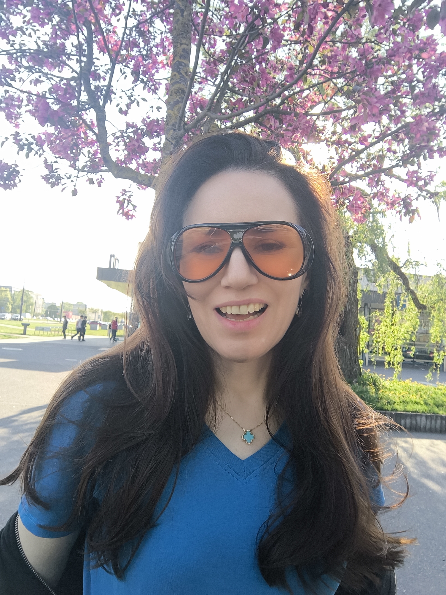 A selfie of a woman wearing orange-tinted aviator sunglasses and a blue V-neck t-shirt is taken outdoors. She has long dark hair and a turquoise pendant necklace. Behind her, a tree with pink blossoms is visible. In the background, a park setting can be observed with other trees and some people walking in the distance. A grey paved ground is at the lower part of the shot. Based on the architectural style and general appearance of the surroundings, the location appears to be Riga, Latvia.