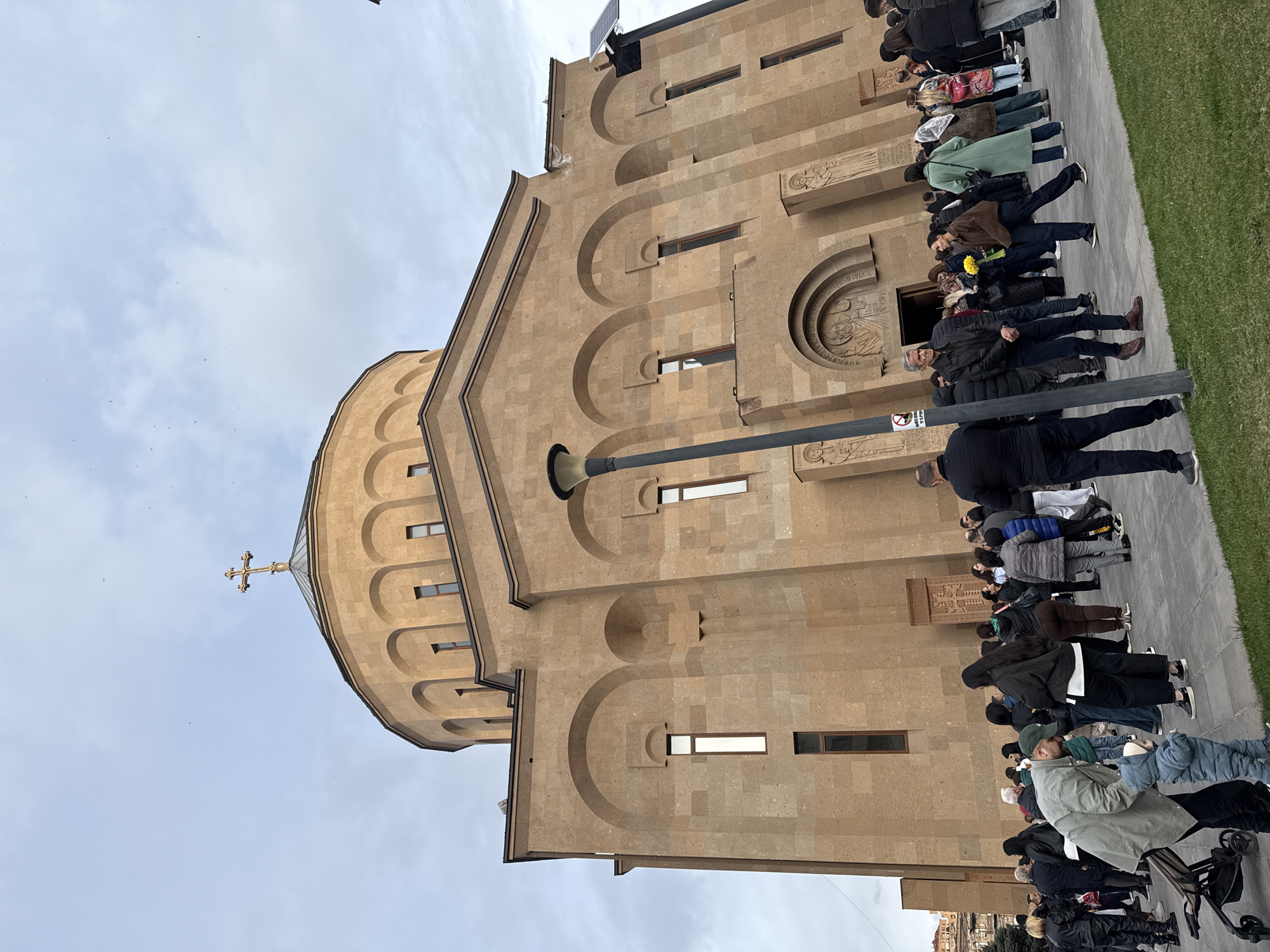 The image shows Saint Anna Church in Yerevan, Armenia. A crowd of people is gathered outside the church. The church is a light brown stone building with a dome topped by a cross. The sky is overcast. The people are standing in a line along the sidewalk. Some appear to be tourists or visitors. There is grass along the edge of the sidewalk. A black metal pole is in the foreground.