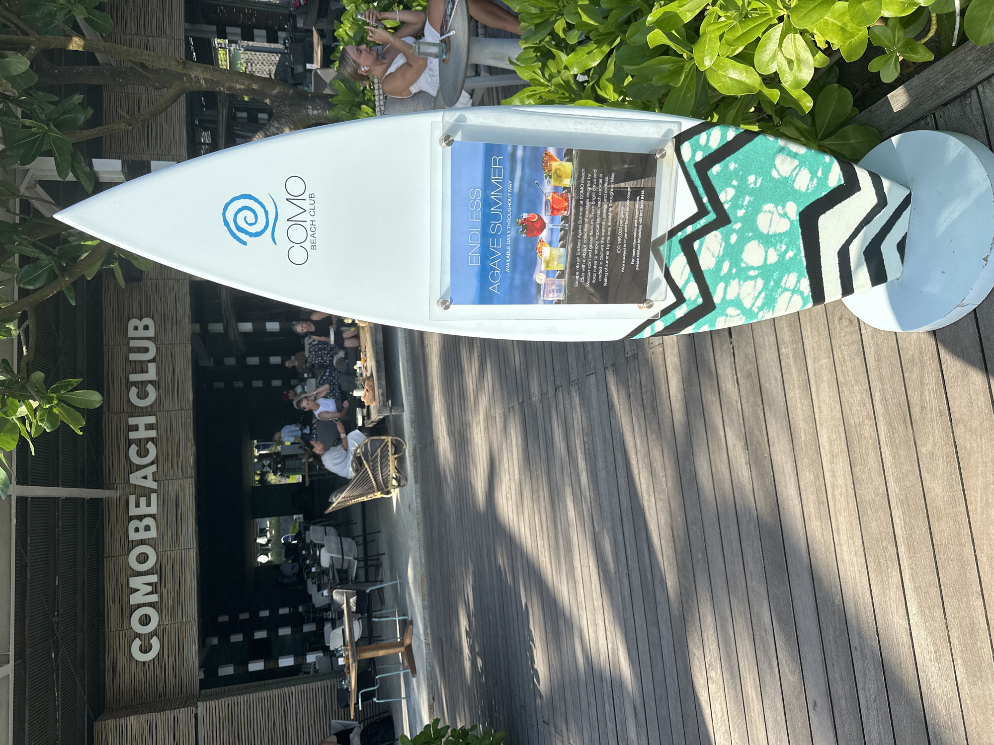 The photograph shows a promotional display for the COMO Beach Club, located in North Kuta, Indonesia. The display resembles a surfboard, with the club's logo and a poster advertising an "Endless Agave Summer" cocktail promotion available daily throughout May. The poster features multiple cocktail glasses with brightly colored alcoholic drinks.
The background reveals a partially obscured view of the interior of the COMO Beach Club, with people seated at tables. The flooring of the area around the promotional sign is made of wooden planks. A section of green foliage can be seen behind the sign.
One person is visible sitting at a table near the sign, with a second person in the background on the same table.