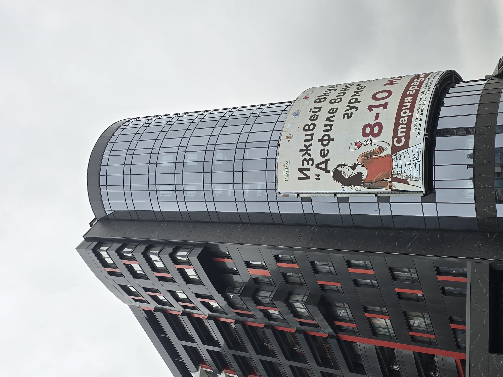A tilted, low-angle shot captures a modern building with a distinctive architectural design against an overcast sky. The upper section of the building features a curved, glass-paneled facade, reflecting the cloudy sky. Attached to this curved section is a large, vertical banner advertising an event. Below the glass section, the building transitions into a darker, more angular facade with geometric patterns, black panels, and horizontal red accents highlighting the window frames.
The banner prominently displays text in Cyrillic, along with a stylized illustration of a woman in a red sweater, lying down and holding a wine glass. The text, translated from Bulgarian, reads: "Plovdiv," "Experience taste," "'Wine Gourmet Defile'," "8-10 M" (likely May), "Old Town," "Tourist festival 'Wine Defile'," and partially, "The annual program for development as...". The overall advertisement points to a wine and gourmet festival happening in Plovdiv's Old Town.
The weather appears to be overcast, indicated by the uniformly grey sky. The unique angles and contrasting materials of the building create a striking, contemporary urban scene.