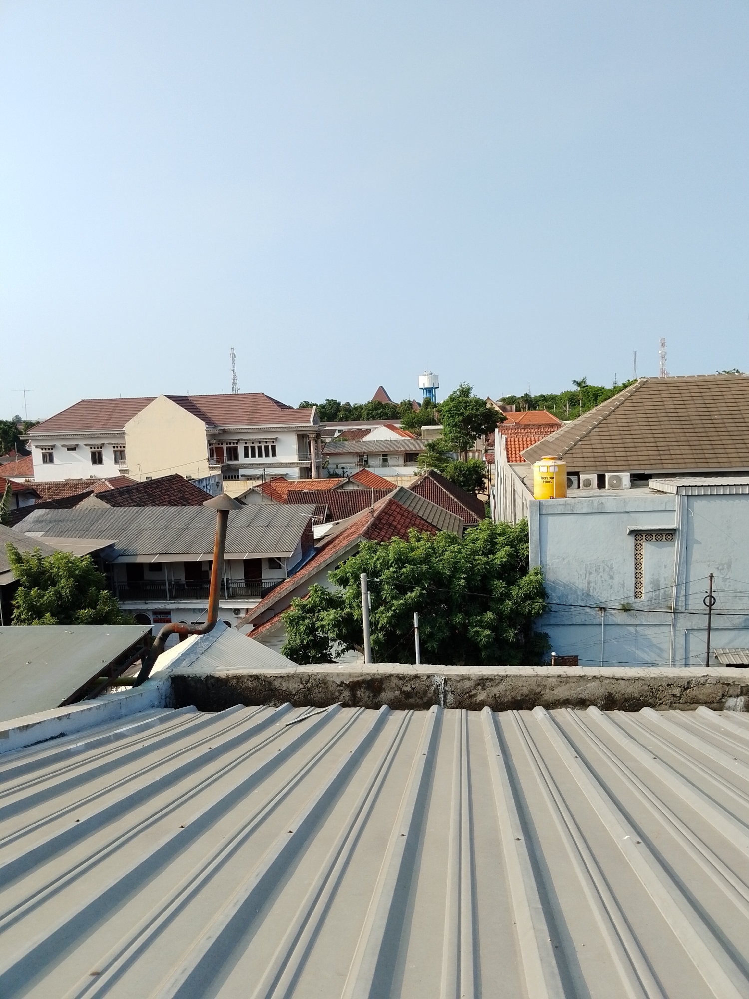 The image displays a panoramic view of rooftops in a residential area of Pasuruan, Indonesia. The foreground is dominated by a corrugated metal roof, suggesting a modern construction or a renovation. Beyond this, a cluster of houses with terracotta tiled roofs and white-walled buildings are visible, interspersed with lush green trees. A prominent yellow water tank sits atop a light blue building in the middle ground, alongside air conditioning units. In the distance, communication towers and a water tower indicate the urban setting. The sky is clear and blue, suggesting a sunny day. The architectural styles vary, hinting at a blend of traditional and contemporary housing in this part of Pasuruan. There are no people or animals visible in the scene.