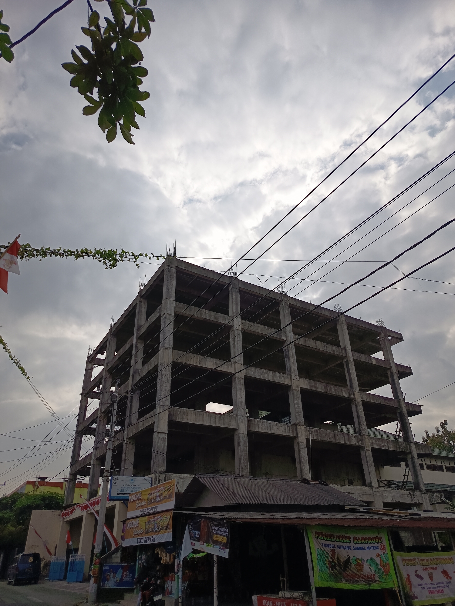 The image depicts an unfinished concrete building under construction, likely in the city of Ngaglik, Indonesia, due to the presence of Indonesian flags. The building has multiple stories, each with exposed concrete floors and columns. Overhead power lines traverse the sky, with some branches of a tree extending into the upper left corner of the frame. In the foreground, a small street-level structure, possibly a shop or food stall, is visible. This structure has a corrugated metal roof and displays various colorful banners, some featuring text in Indonesian, including one advertising "PECEL LELE SARIMOSE" and another with "INDOMIE". A dark blue van is parked on the left side of the street, and a motorcycle is partially visible in the lower right. The sky is overcast with visible clouds. The scene appears to be captured during daylight hours.