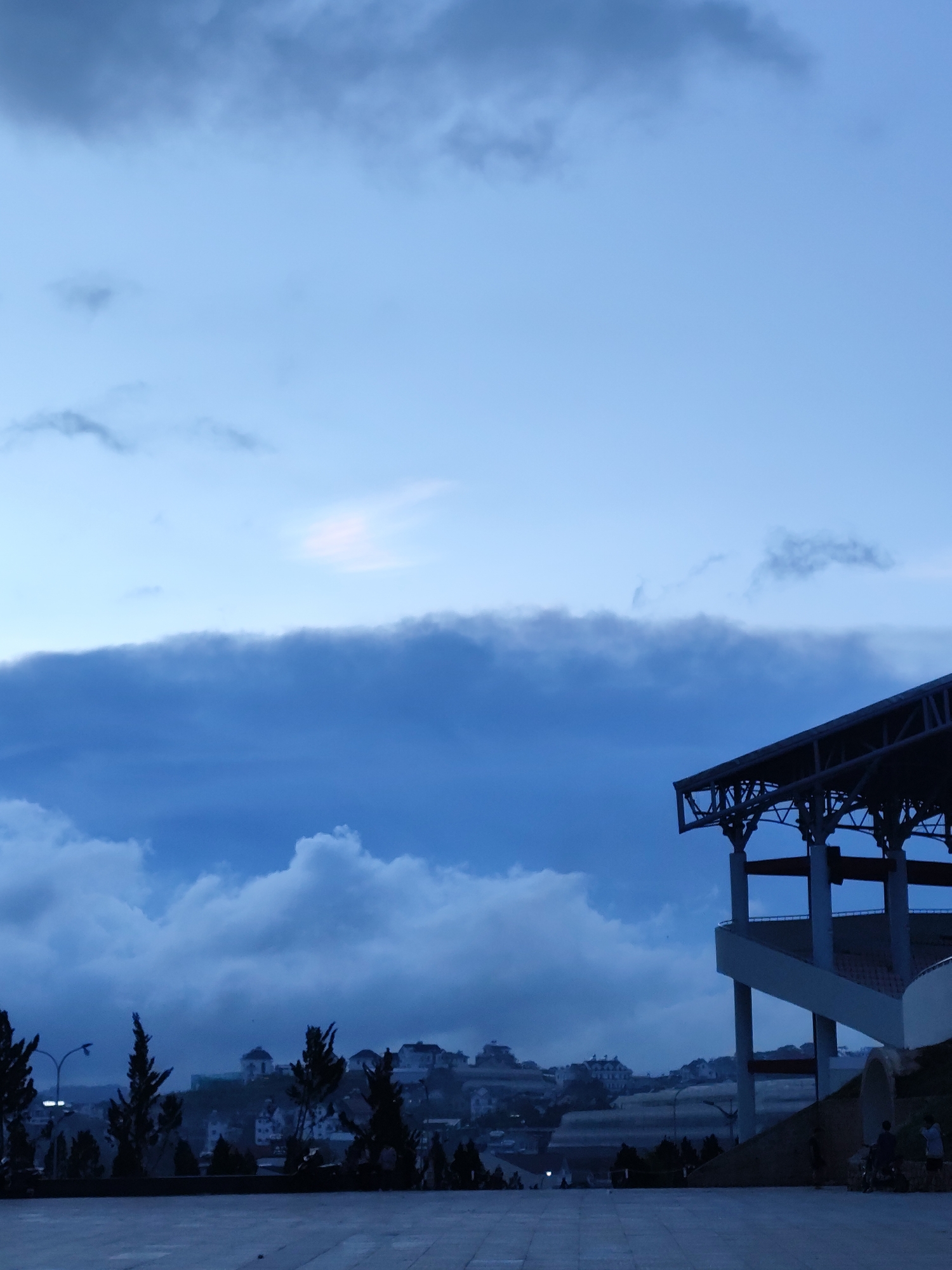 The image depicts a landscape in Lang Biang - Da Lat, Vietnam, under an overcast sky. The scene features a modern stadium-like structure with visible steel supports and concrete tiers. In the background, a cityscape with densely packed buildings is visible, silhouetted against the sky. Trees are scattered throughout the foreground and midground. A paved area extends into the foreground, and several individuals are visible in the lower right, suggesting pedestrian activity. The overall lighting is subdued, casting the scene in shades of blue and grey.