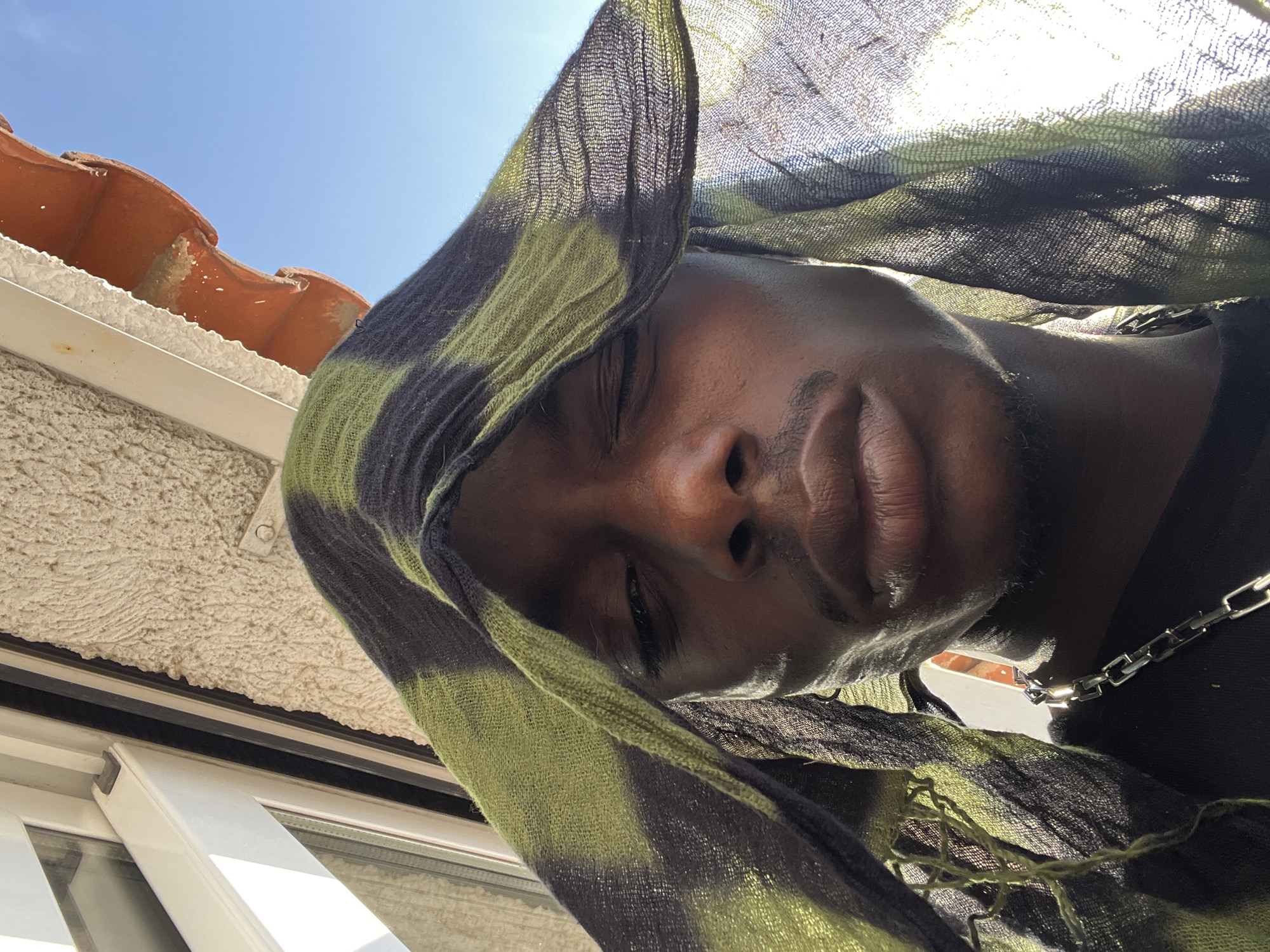 A close-up, low-angle shot depicts a Black person with closed eyes. They are draped with a loosely woven scarf, patterned in shades of green and black. The light of the sun illuminates the fine texture of the fabric and the person's face. Visible above and to the side of the person are elements of architecture: a cream-colored, textured wall; a horizontal, white window frame; and red roof tiles. A section of a metal chain necklace is also visible around the person's neck. The architectural details indicate a Mediterranean context, likely within the city of Barcelona, Spain, due to the typical building styles found there.