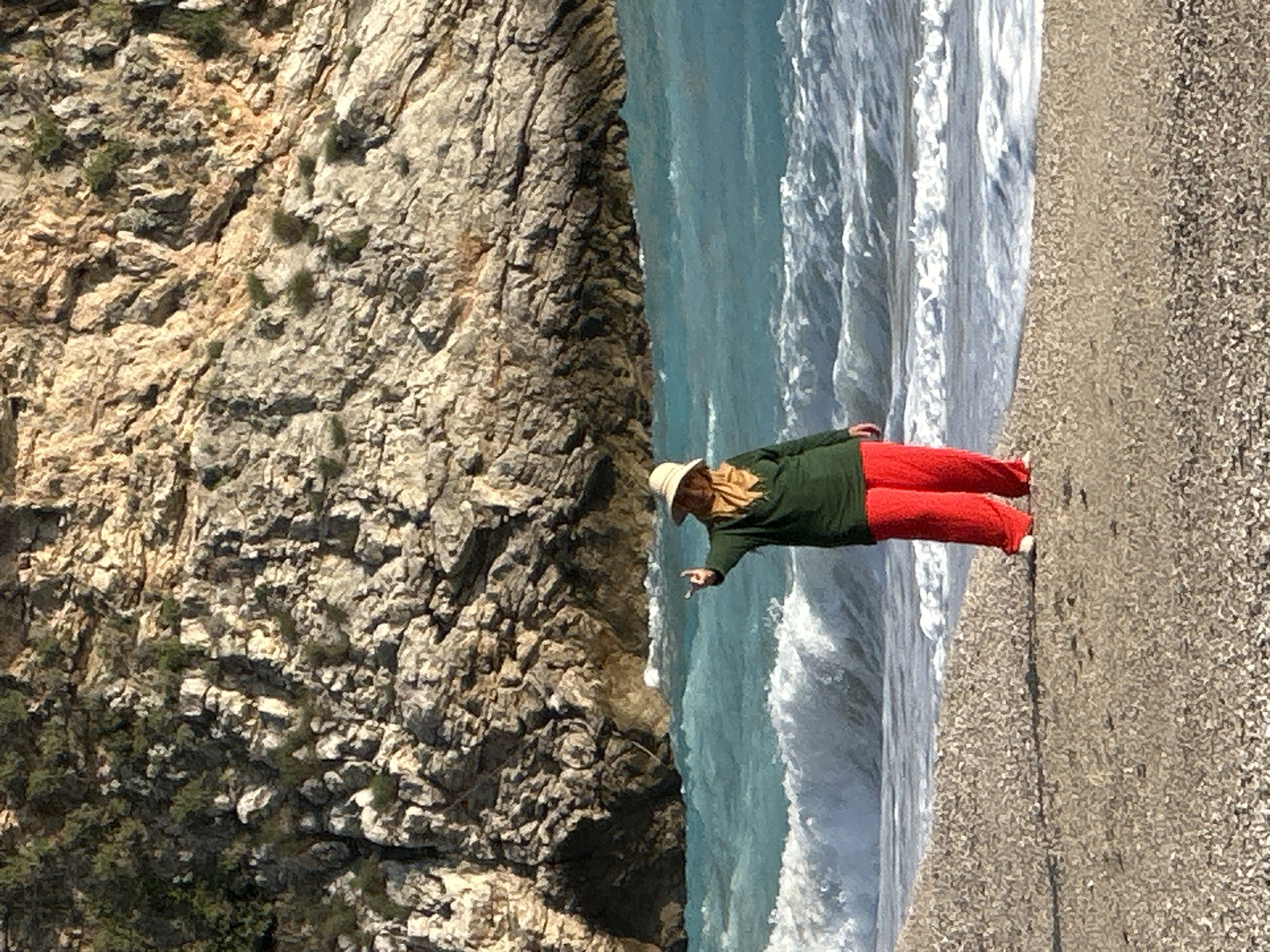 A vertical, color photograph shows a beach scene. An adult person wearing a tan hat, dark green long-sleeve shirt, and bright red pants stands on a light gray beach, partially in the surf. The person is pointing towards a large, tan-gray rock face on the left. The water is turquoise, with white surf. The beach is light gray, composed of small stones. The person may be located in Fethiye, Turkey.