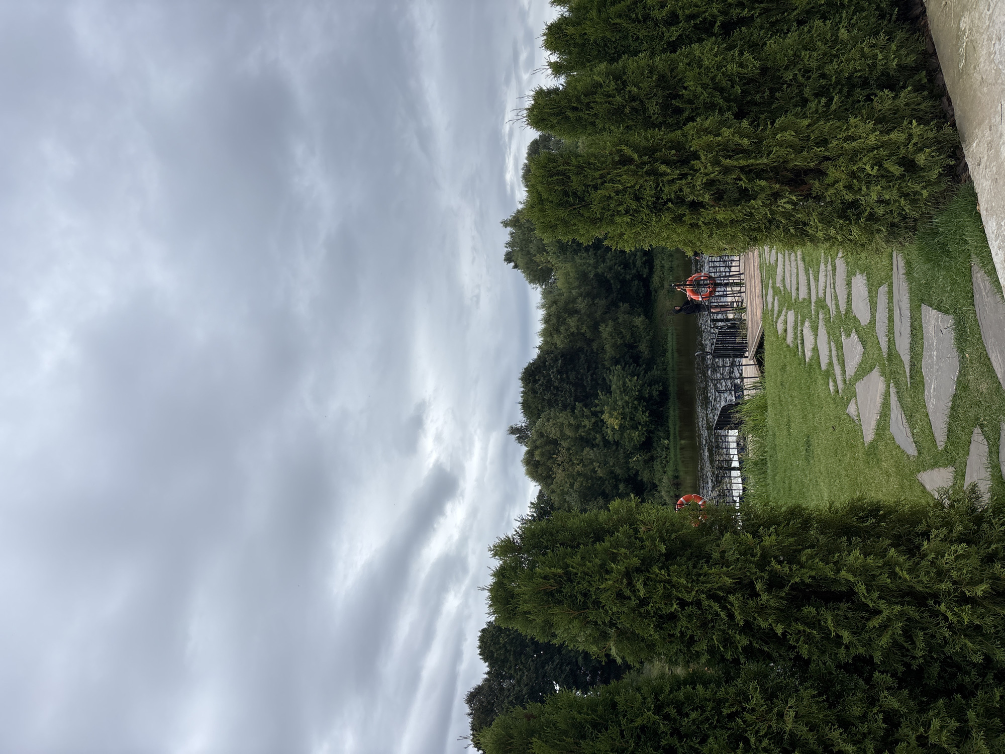 The image captures an overcast day with a cloudy sky over a body of water and a verdant landscape. In the foreground, several tall, green evergreen trees stand symmetrically, framing a stone pathway made of irregularly shaped gray flagstones laid on green grass. The pathway leads towards a wooden dock on the water.
On the dock, two individuals are visible. One, wearing dark clothing and a hood, appears to be standing. Near them, an orange life preserver is attached to a railing. A second person, also in dark clothing, is standing further back on the dock. The water is dark and appears calm, reflecting the muted tones of the sky.
Behind the water, a dense line of trees forms a backdrop, suggesting a natural or park-like setting. The overall atmosphere is serene and naturalistic. The location is a pier or a waterfront area in the city of Zvenigorod, Russia.