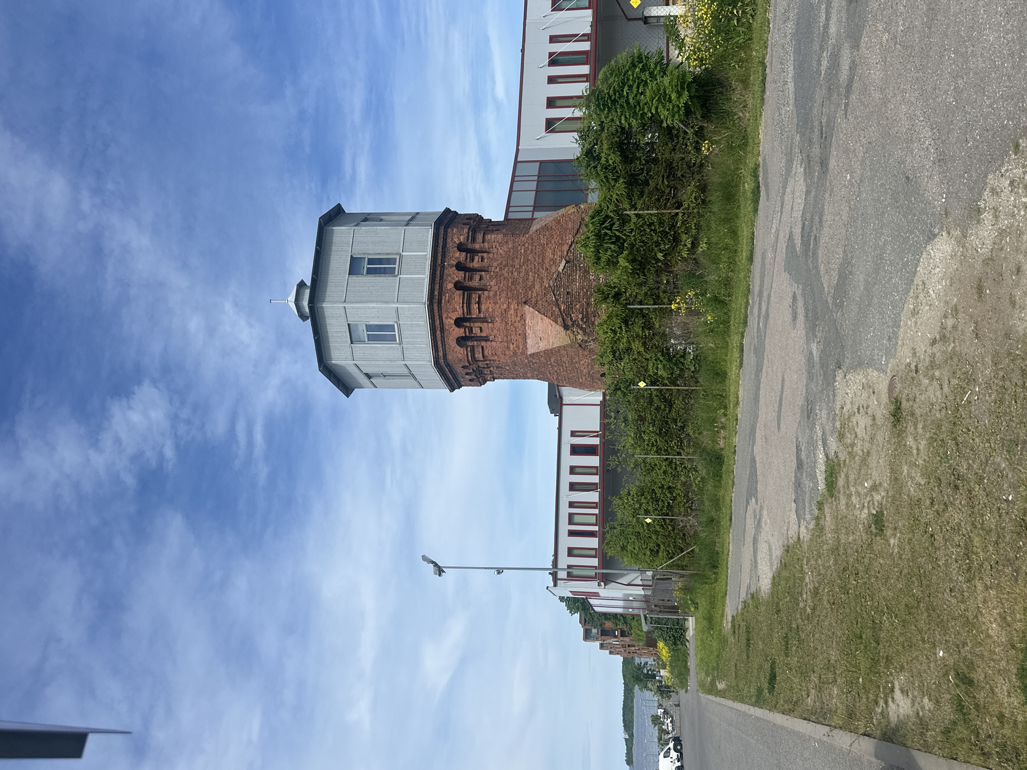 The image depicts an exterior view of a historical brick water tower adjacent to modern buildings in Tallinn, Estonia. The water tower, constructed of red brick with a distinctive octagonal grey metal roof, features arched detailing near its top. It is integrated with a contemporary building featuring a white facade, large glass windows, and red window frames. The scene is set on a sunny day with a blue sky dotted with white clouds. In the foreground, a paved road runs alongside a grassy verge. Behind the fence in the midground, there is lush green foliage and yellow wildflowers. In the background, to the left, a body of water is visible, with a distant shoreline and buildings. Several white vans are parked along the roadside further down the street, suggesting this is an industrial or commercial area. No people are visible in the frame.