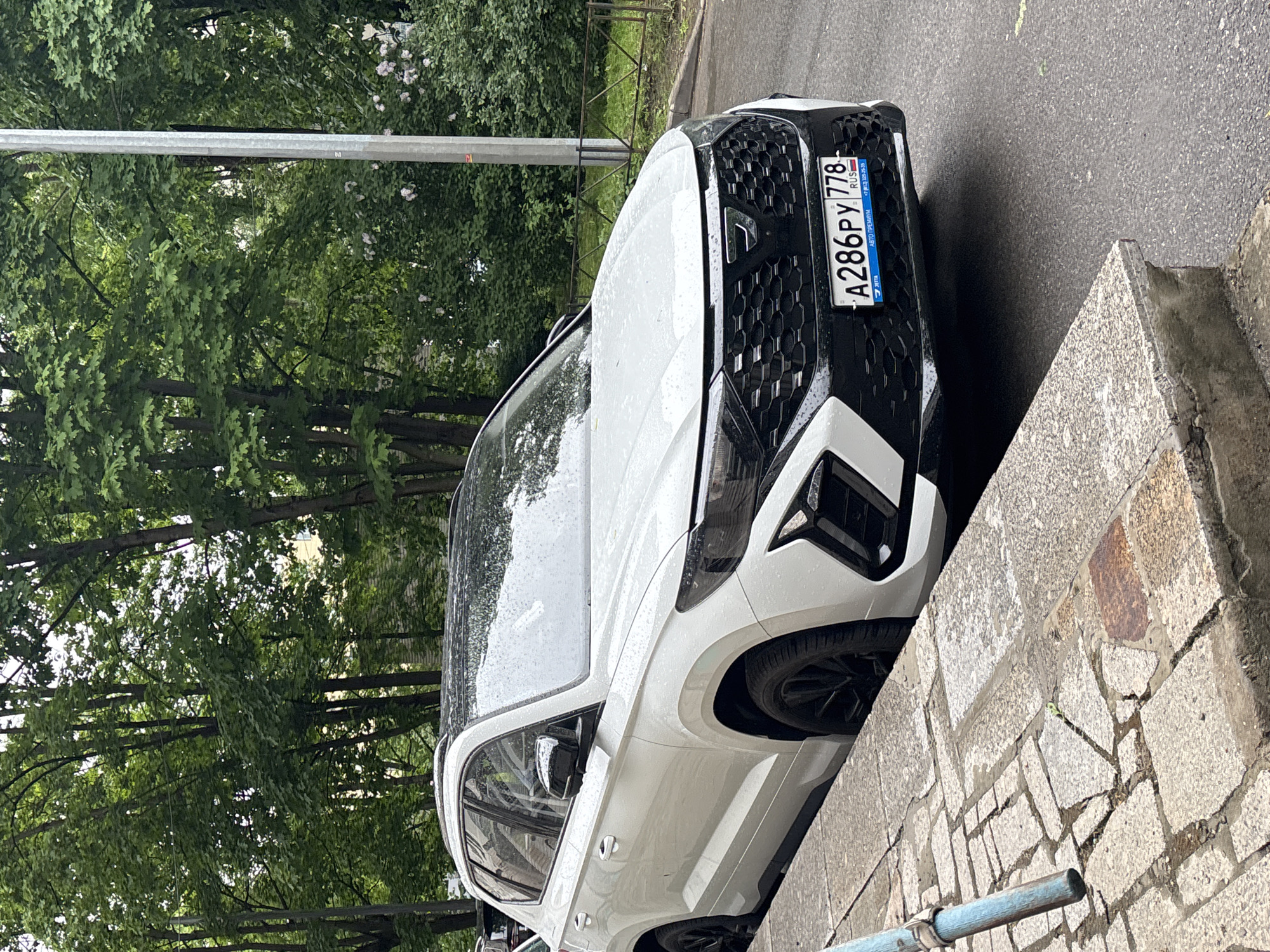 The image shows a white Cupra Formentor parked on a street in what appears to be Saint Petersburg, Russia. The vehicle is partially on the sidewalk, next to a stone retaining wall. The license plate is visible and reads "A286PY 778", indicating it is registered in Russia. The surroundings include green trees and a metal pole. The weather seems to be overcast or after a recent rain. No people are visible.