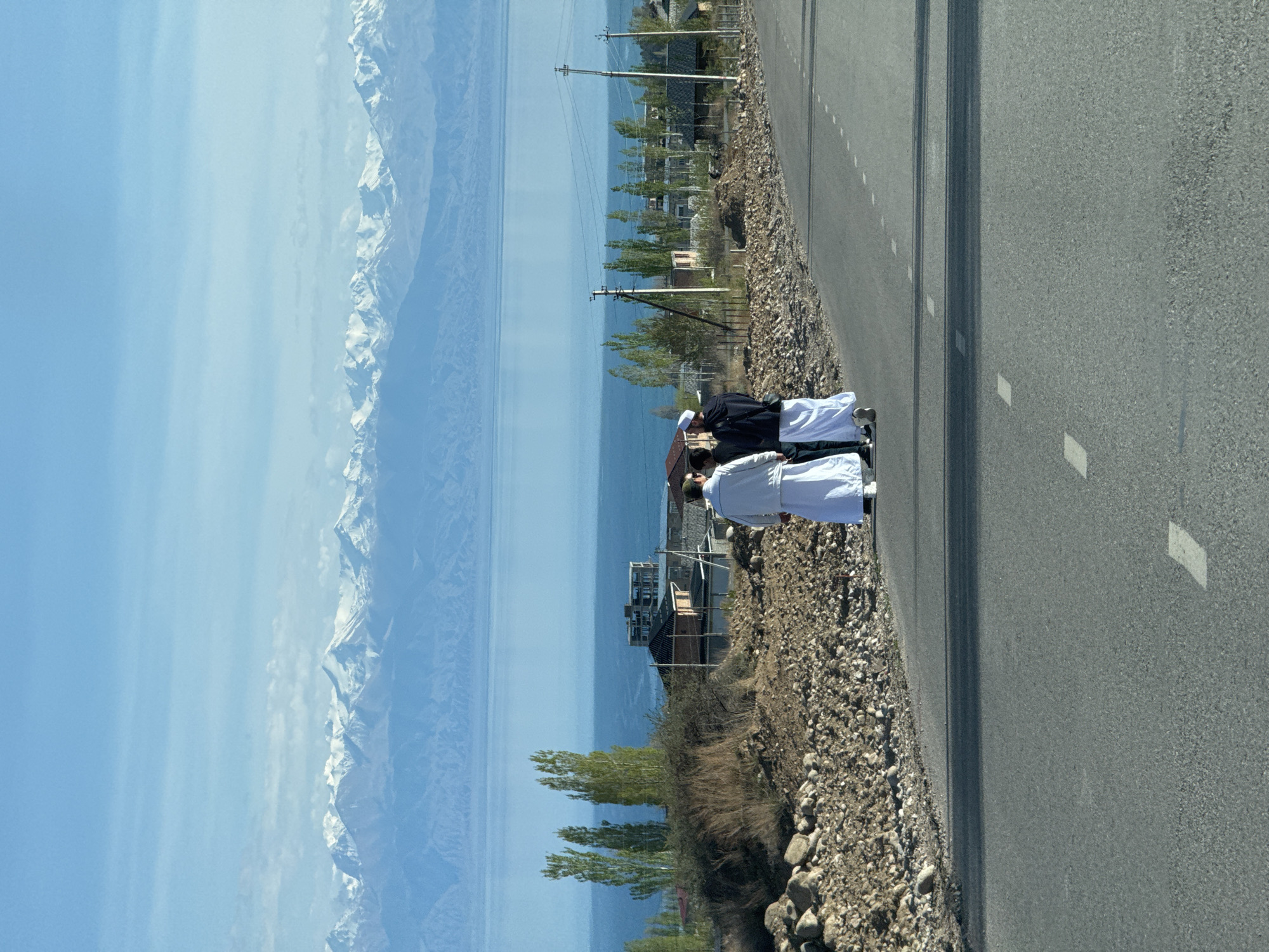 The image shows a scene along a road in Çolpon-Ata, Kyrgyzstan. A group of people are standing on the side of the road. Two individuals are wearing white robes and hats, while another is wearing a black jacket. The road is paved with asphalt and has white lines marking the lanes. Beside the road is a rocky embankment with some vegetation. In the background, there is a body of water (likely a lake), and beyond that, snow-capped mountains are visible under a clear blue sky. Utility poles and some buildings are also present along the lakeshore.