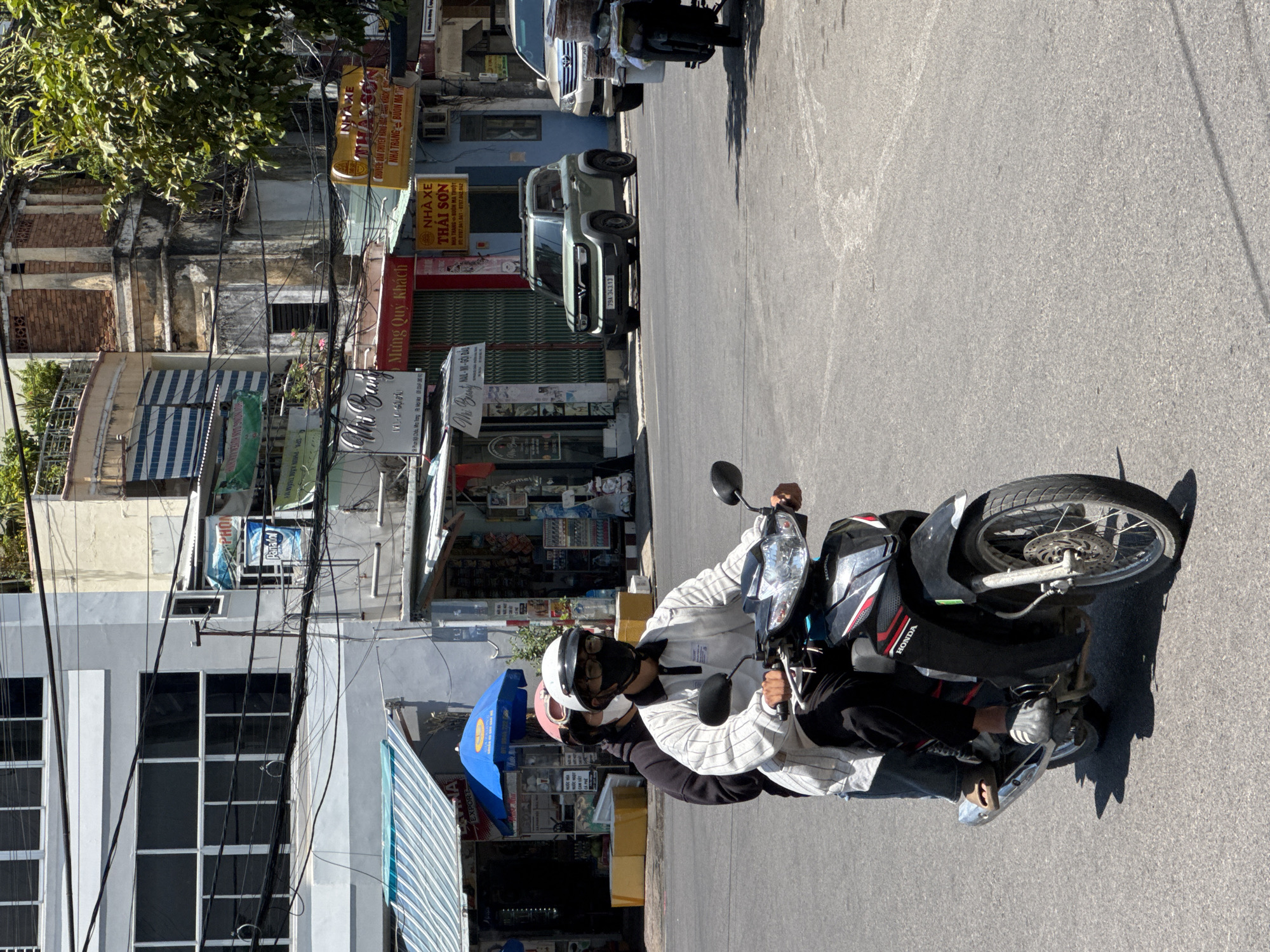 The image depicts a street scene in Nha Trang, Vietnam, under bright daylight. In the foreground, two individuals are riding a black Honda scooter. The rider in front wears glasses, a white and grey pinstriped collared shirt, and a white helmet with a black face mask. The passenger behind wears a black hoodie and a pink helmet. They are positioned on the right side of the road, moving forward.
The background reveals a bustling urban environment with several buildings. On the left, a light grey building with large, dark-paned windows dominates the upper portion. Below it are storefronts with blue and white striped awnings. Further to the right, older buildings with exposed brick and weathered facades are visible, interspersed with signs and utility wires. One sign, in yellow and red, reads "NHÀ XE THÁI SƠN". A light green SUV is parked on the right side of the road, and a light-colored SUV is visible behind it. The street surface is asphalt, with some tire marks and shadows cast by the overhead sun. The overall atmosphere is one of a typical day in a Vietnamese city, with traffic and commercial activity.