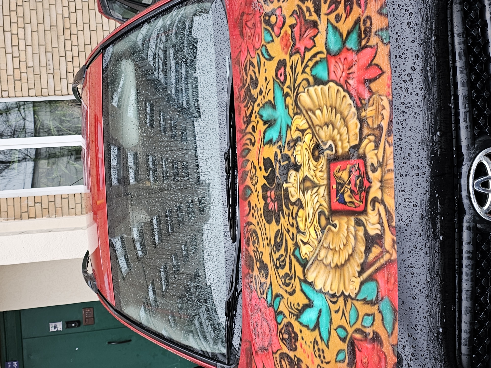 The image depicts a red car parked in Khimki, Russia. The car's hood is adorned with a colorful, folk-art style painting featuring floral patterns, a golden double-headed eagle, and a square emblem. The windshield is wet, reflecting a nearby building with a brick facade and white-framed windows. The car is parked near a green door with a keypad and intercom system. The front of the car, including the grill with a Toyota logo, is visible and covered in raindrops.