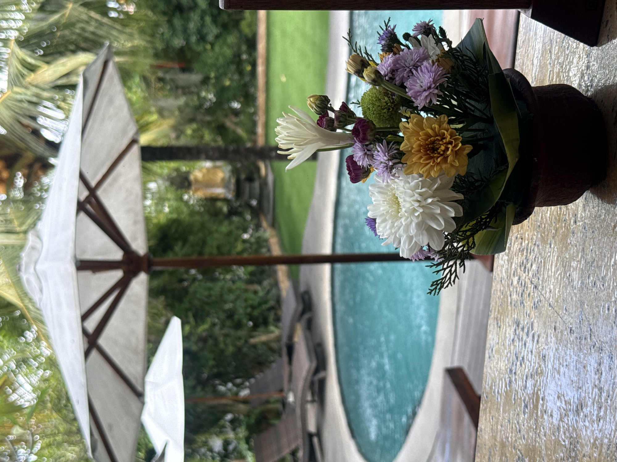 A flower arrangement in a brown pot rests on a textured, light-colored surface, possibly a table or ledge. The arrangement features a mix of white, yellow, and purple flowers, along with green foliage. In the background, there is a turquoise-colored swimming pool, surrounded by green grass and lush trees. A white umbrella with a brown frame is also visible, partially obscuring the view of the background. The scene is possibly located in or near North Kuta, Indonesia, given the tropical vegetation. The arrangement is in the foreground. The environment suggests a tropical resort or private residence.