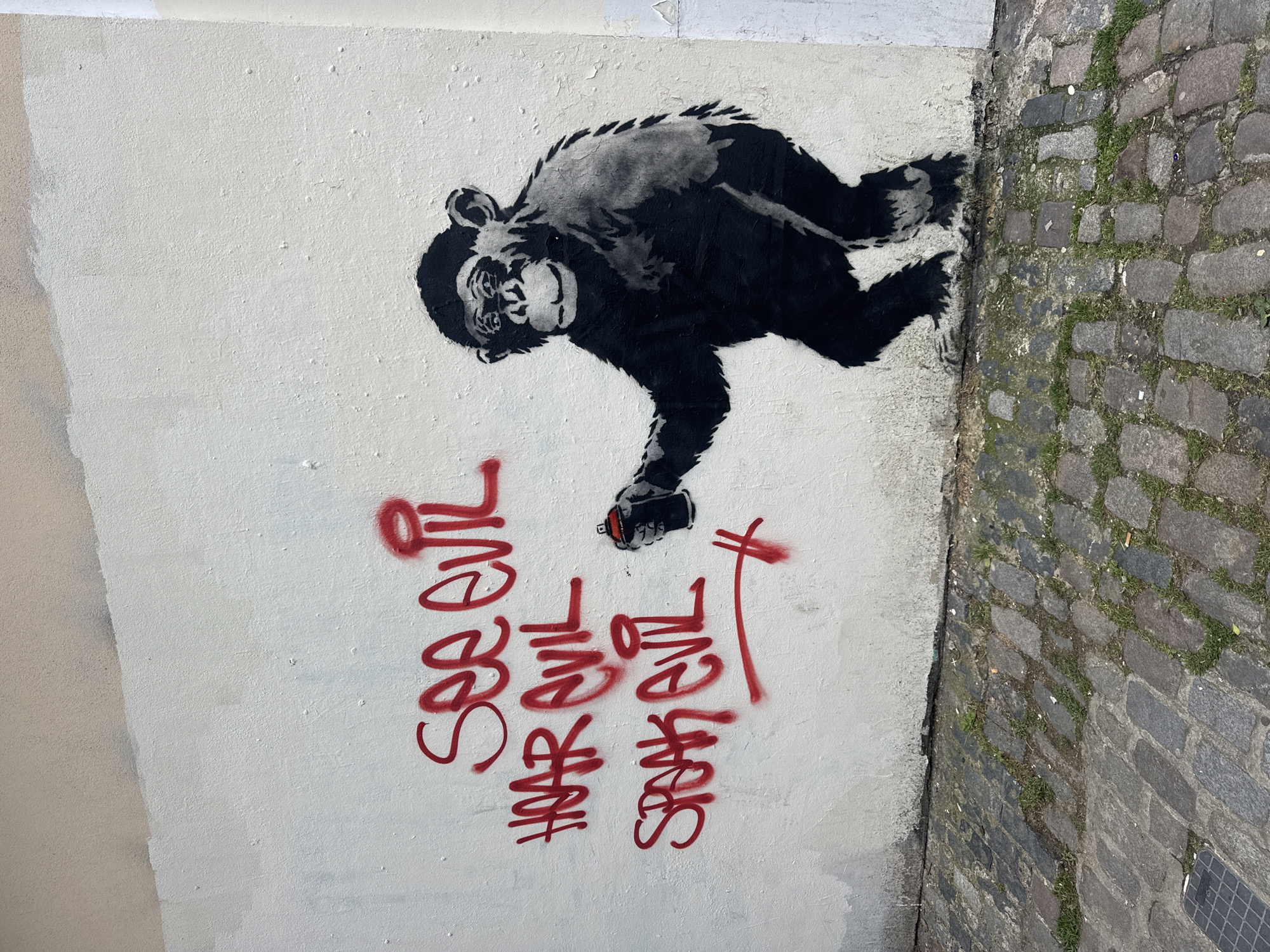A street-level photograph shows a mural on a white wall next to a cobblestone street. The mural features a stenciled chimpanzee, predominantly black and white, with a spray can in its hand. Below the chimpanzee, in red spray paint, is the phrase "See Evil" repeated three times, each written in a unique, stylized font. The wall's surface is slightly textured, while the cobblestone street to the right exhibits signs of wear and vegetation. The image captures a juxtaposition of street art and urban infrastructure. Based on style and subject matter, the mural is likely attributed to the artist Banksy. The location is in London, United Kingdom.