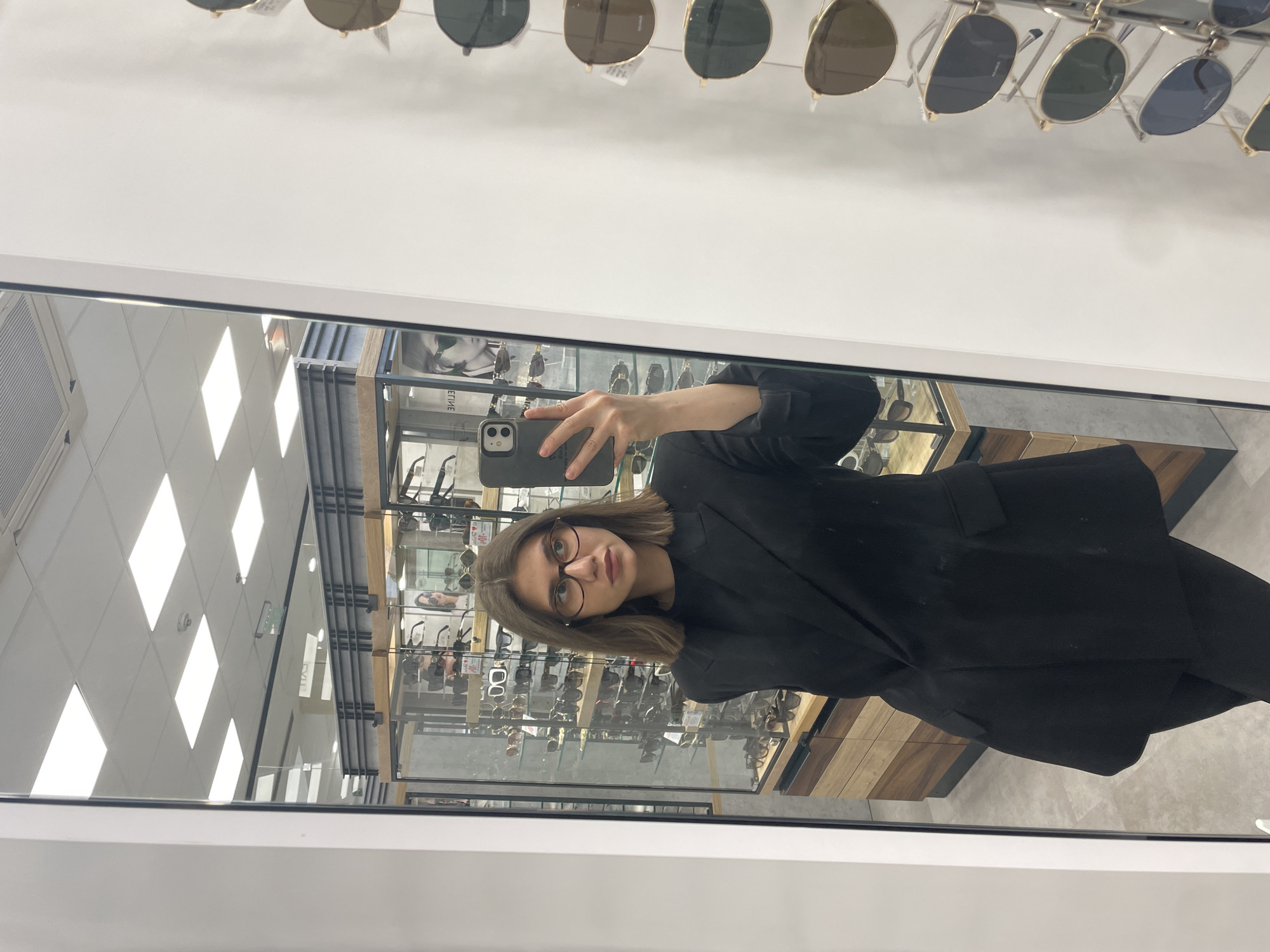 A woman in a black blazer and glasses holds up a cell phone to take a selfie in a mirror in what appears to be a store. Behind her in the mirror is a display case containing eyeglasses. Above her in the frame are sunglasses hanging on a rack. The environment suggests an optical shop or department store. The location is unknown.