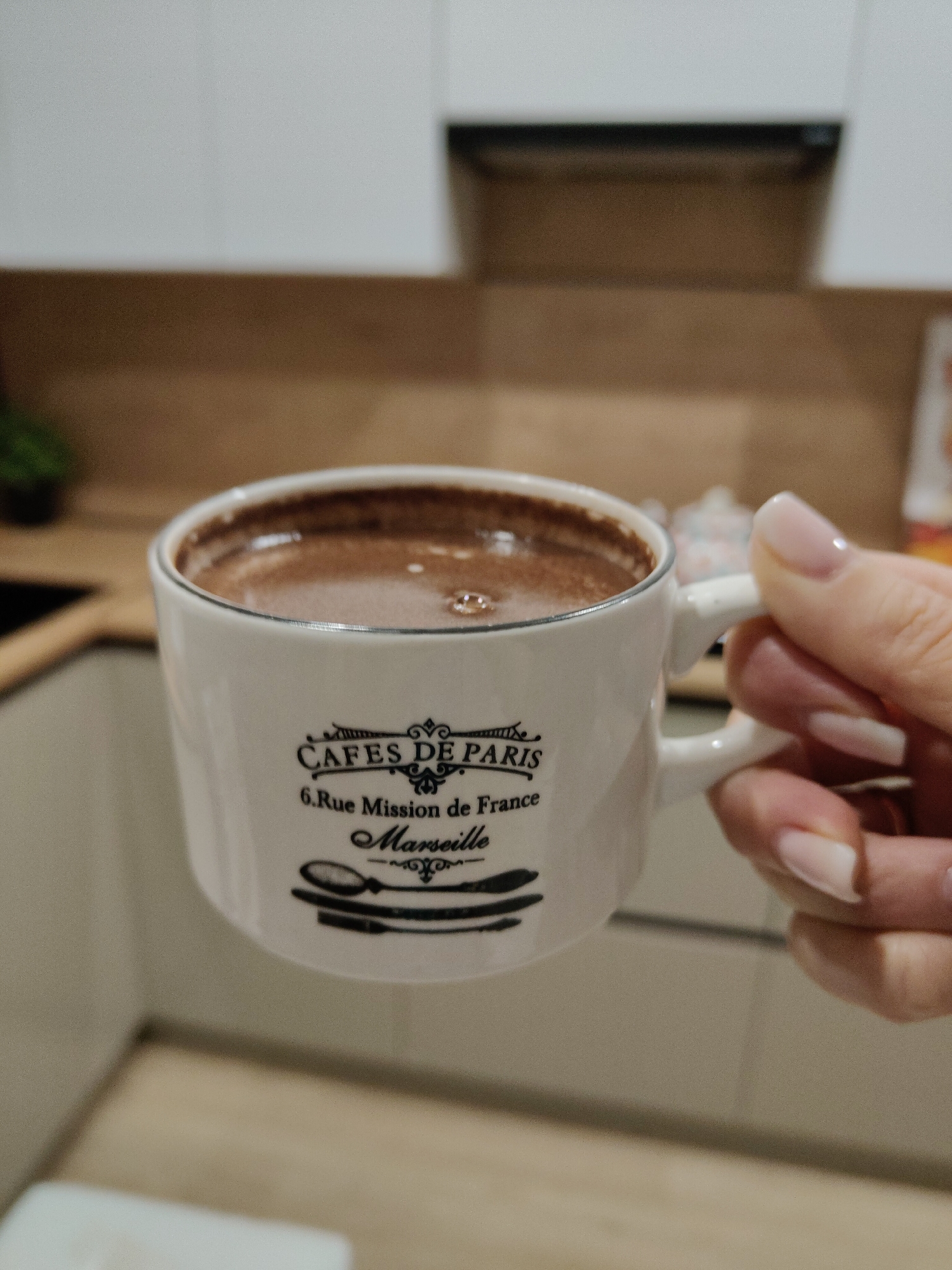 A person is holding a white mug with the words "CAFES DE PARIS" and "6.Rue Mission de France Marseille" printed on it. The mug is filled with a brown liquid, likely hot chocolate or coffee. The background shows a kitchen setting with light wood countertops and white cabinets. The location is Unknown, Unknown.