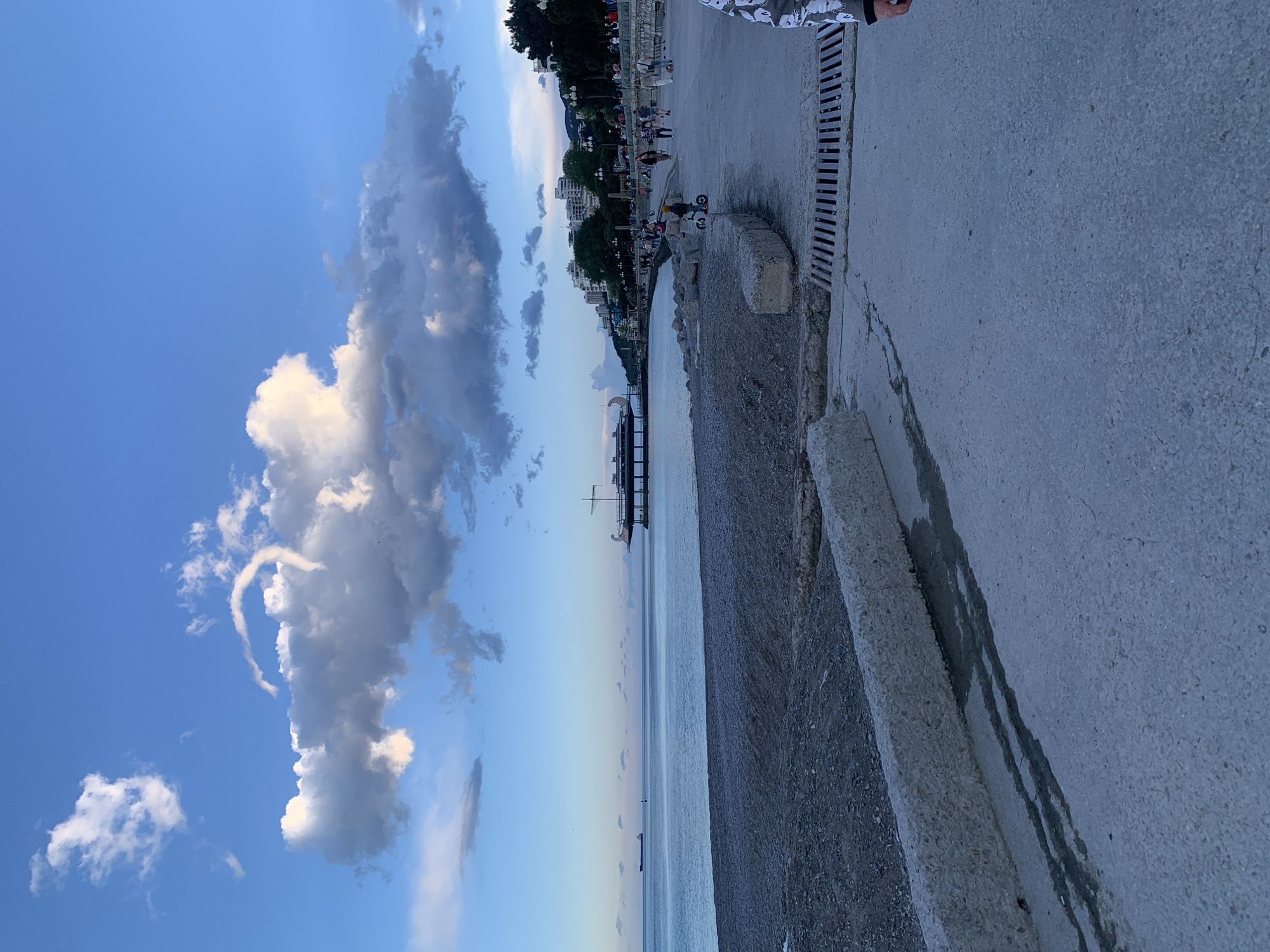 The image captures a coastal scene. On the right side, a concrete promenade with a drainage grate along the edge is visible. A pebble beach runs parallel to the promenade, bordered by concrete blocks. In the background, a city skyline with buildings and trees stretches along the coast. A pier extends into the sea. The sky is a gradient of blue with scattered clouds, some of which form a distinctive ring-like shape. People are walking along the promenade.