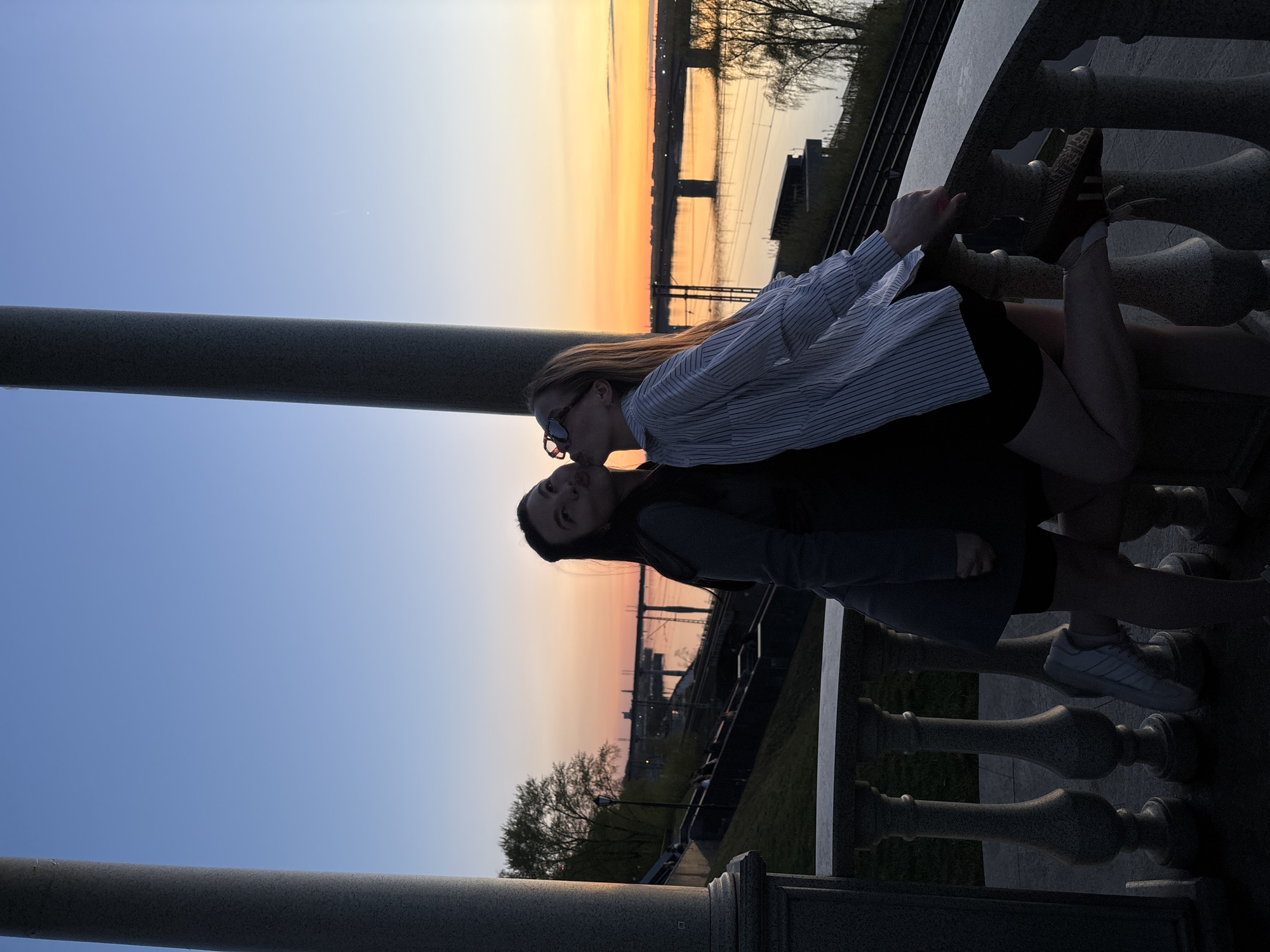 Two young women are posing for a photograph on a stone balustrade at sunset. The woman in the foreground wears a dark jacket and black shorts. The woman behind her wears a striped button-down shirt, black shorts, and sunglasses. They are facing each other and appear to be kissing. The background reveals a cityscape and a bridge over a river at sunset. The location is Perm, Russia. A gray pole is visible on the left side of the frame. The balustrade has decorative posts.