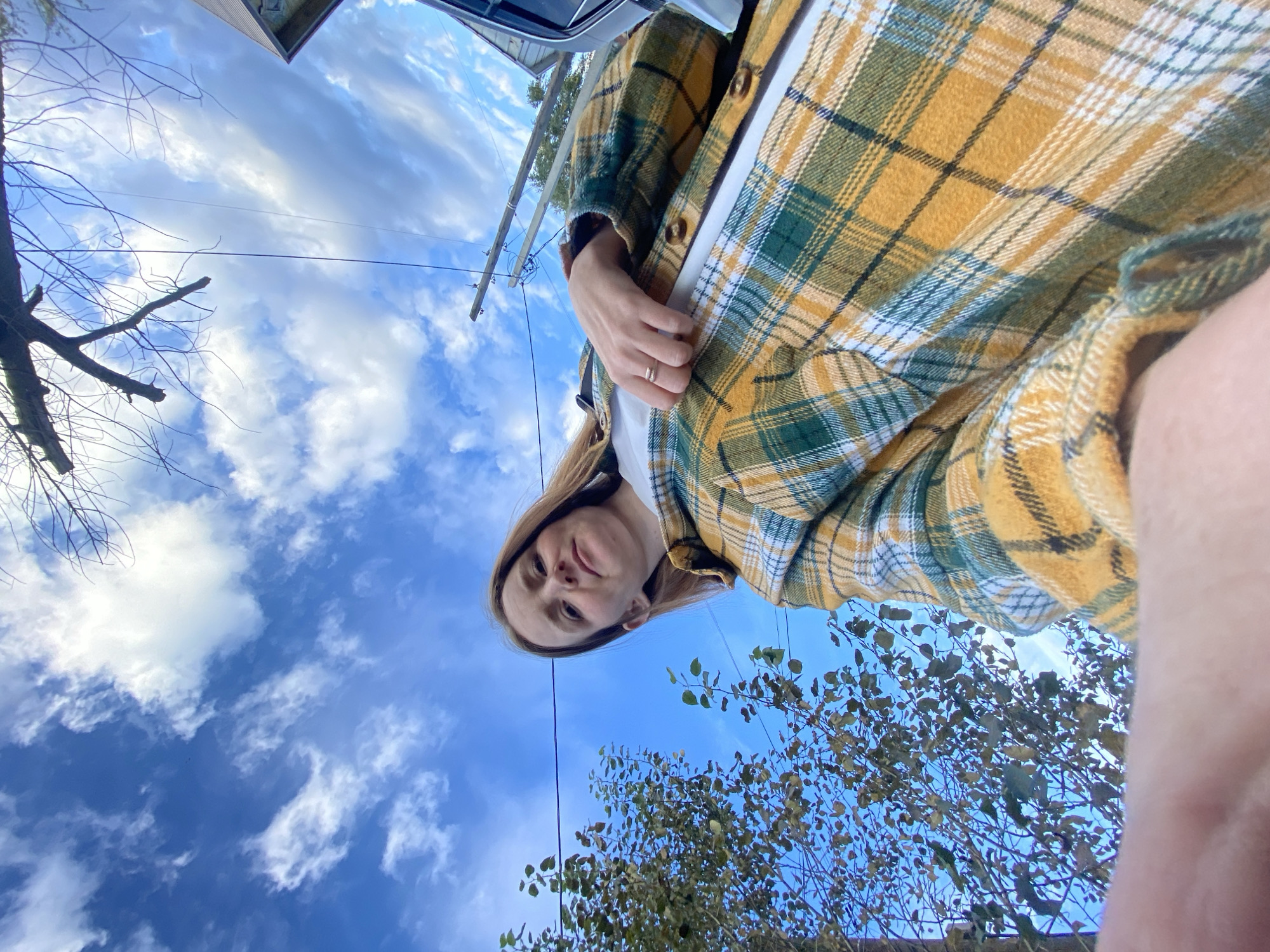 The image is a low-angle, selfie-style shot of a young woman. She has light brown hair and is wearing a white t-shirt and a yellow and green plaid shirt. She is wearing a ring on her right hand. The background is a bright blue sky with white, wispy clouds. To the left of the woman is a tree with sparse, autumnal leaves. There are also power lines visible in the sky. To the right of the woman, part of a vehicle is visible. The image appears to have been taken outdoors. The location is specified as Chaikovsky, Russia.