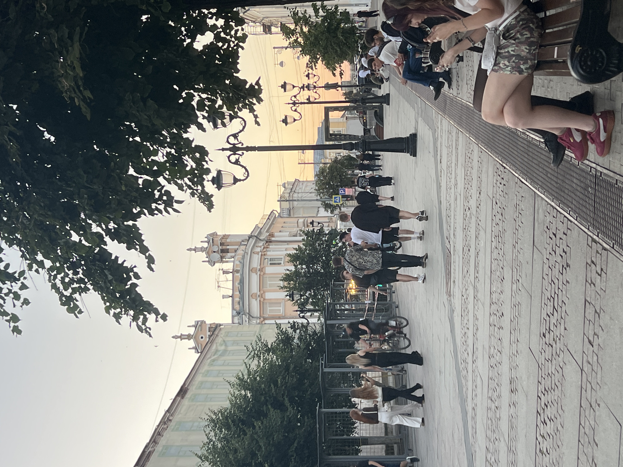 The image depicts a street scene in Tver, Russia, with a pedestrian walkway paved with decorative stonework. Several groups of people are visible. On the left, three women walk together, two in white tops and one in black. A man on a bicycle is in motion in the middle ground, with a group of men walking nearby. Further ahead, more individuals are strolling and some are seated on benches on the right side of the walkway. Street lamps with ornate designs line the walkway. The background features buildings with classical architectural elements, suggesting an urban environment. The sky is overcast with a soft, warm light, indicating it might be late afternoon or early evening. Trees with dense foliage are present, casting shadows on the pathway.