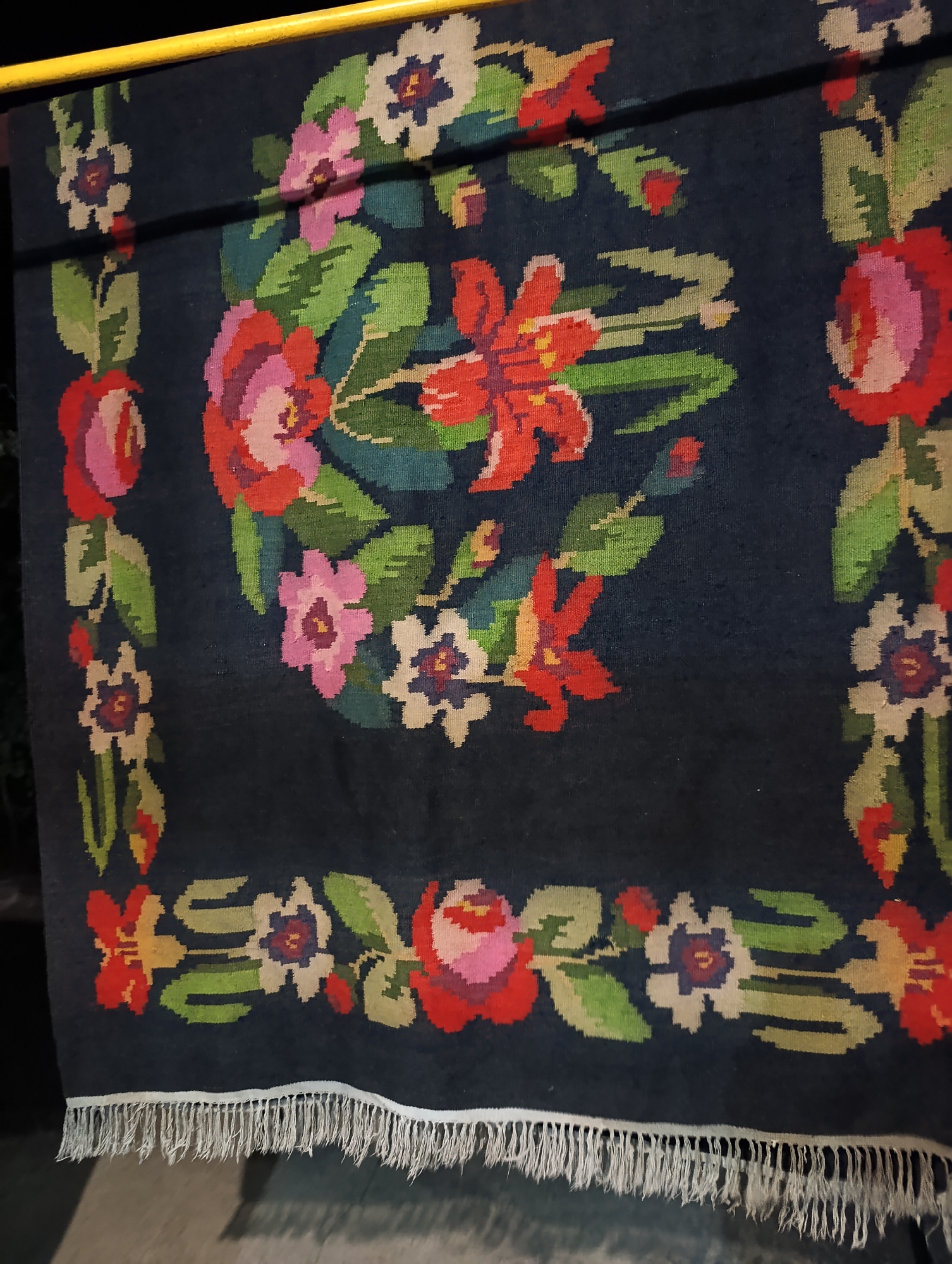 The image shows a close-up of a dark blue rug with a floral pattern, hanging from a yellow bar. The rug is woven with various colors, including red, pink, green, white, and purple, depicting large flowers and leaves. The bottom of the rug has a fringe of white threads. The background is dark and out of focus, with hints of outdoor elements such as greenery and possibly a paved surface. The image was taken in Roghi, Moldova.