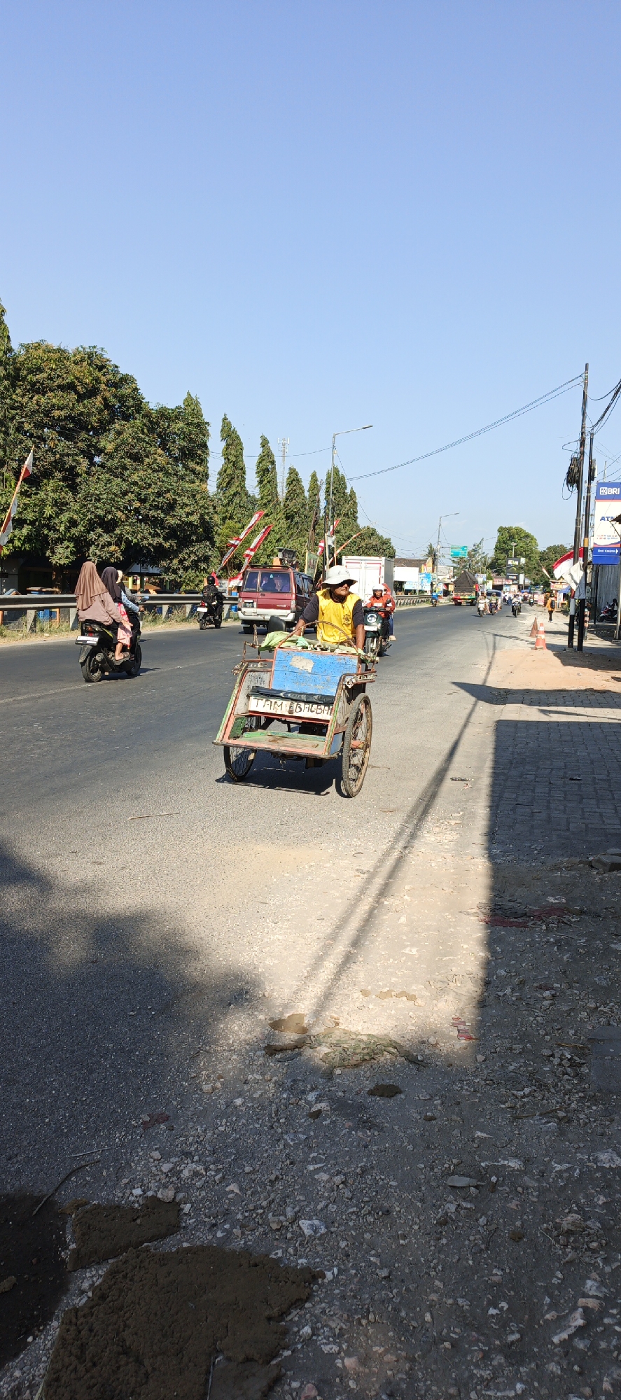 The image depicts a street scene in Kasiyan Timur, Indonesia. A man in a yellow shirt and a hat is operating a pedicab on the right side of the asphalt road. The pedicab has a blue and green body with a license plate that reads "TAM EBBALBAH". Several vehicles, including motorcycles and cars, are visible on the road, with some moving and others parked. Two women on a motorcycle are traveling in the opposite direction to the pedicab. The environment features trees and vegetation along the roadside, with buildings and power lines in the background. The sky is clear and blue, indicating a sunny day. The ground on the right side of the road is a mix of asphalt and dirt, with some debris present.