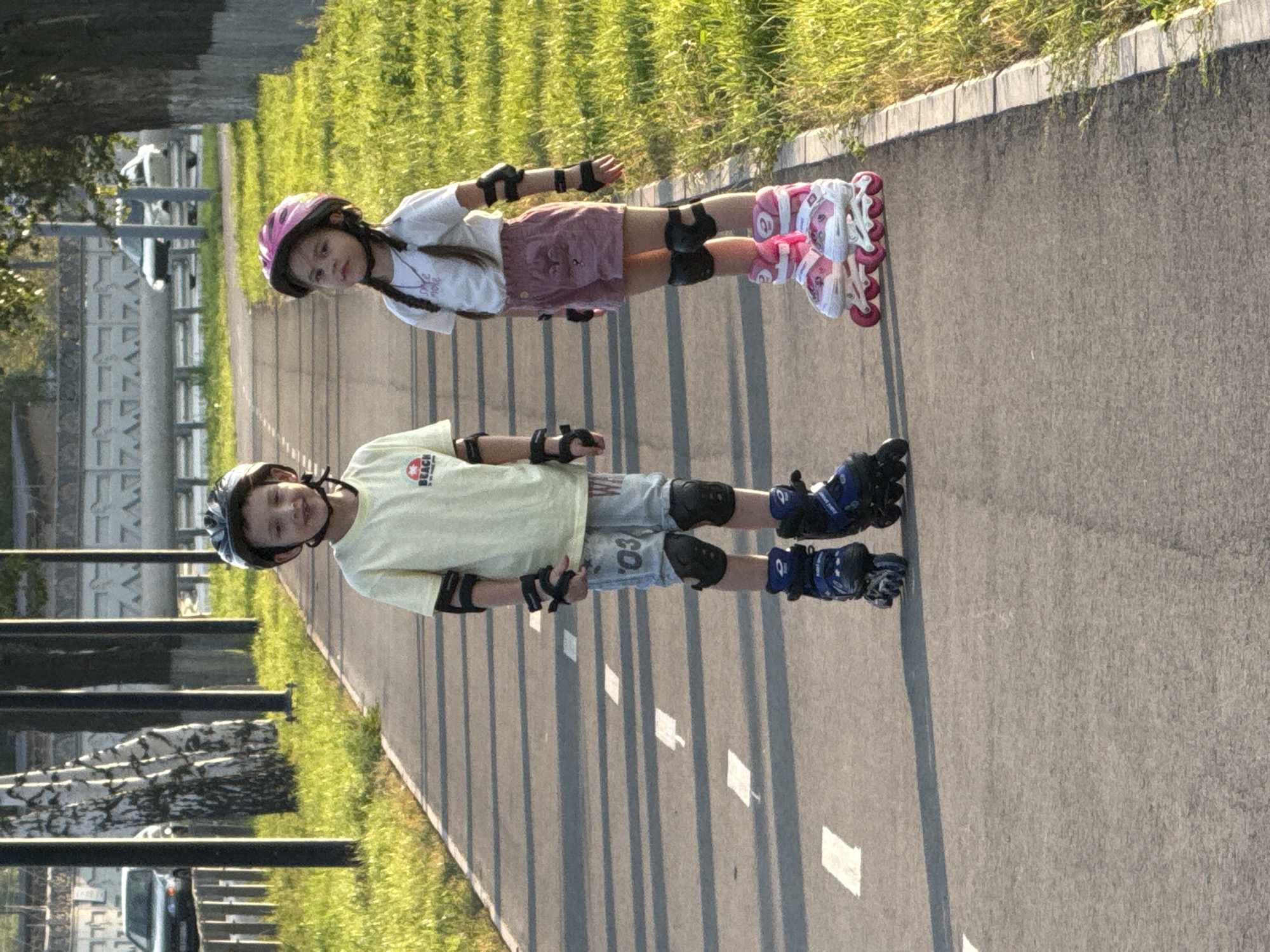 The image depicts two children on rollerblades, positioned vertically in the frame. The child in the foreground, wearing a blue helmet and a light yellow t-shirt, is looking towards the camera with a slight smile. This child is also wearing protective knee and elbow pads, and blue and black rollerblades. The second child, in the background, wears a pink helmet and a white t-shirt with pink shorts. They are also equipped with protective gear and pink rollerblades. Both children appear to be on a paved path, possibly a sidewalk or bike lane, with a grassy area to their left. The scene suggests a sunny day, likely late afternoon or early evening given the long shadows cast on the path. The overall impression is one of outdoor recreation and childhood activity in a park-like or suburban setting. The location context is Votkinsk, Russia. No visible text is discernible on the children's clothing or gear, apart from a small logo on the yellow t-shirt of the foreground child which is too indistinct to read.