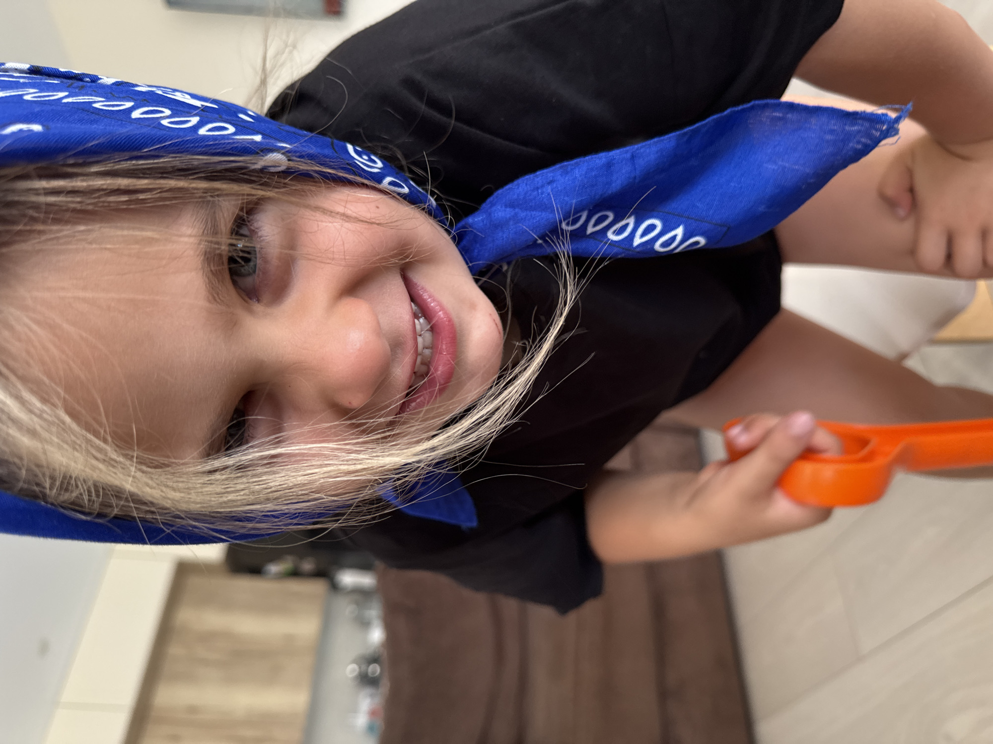 The image is a close-up shot of a young child wearing a blue bandana and a black t-shirt, leaning forward and smiling at the camera. The child's hair is blonde and falls around their face. In their right hand, the child is holding an orange toy shovel. The background is blurred, showing what appears to be an indoor setting with furniture. There is no identifiable information to suggest the location is in Uyutnoye, Ukraine.