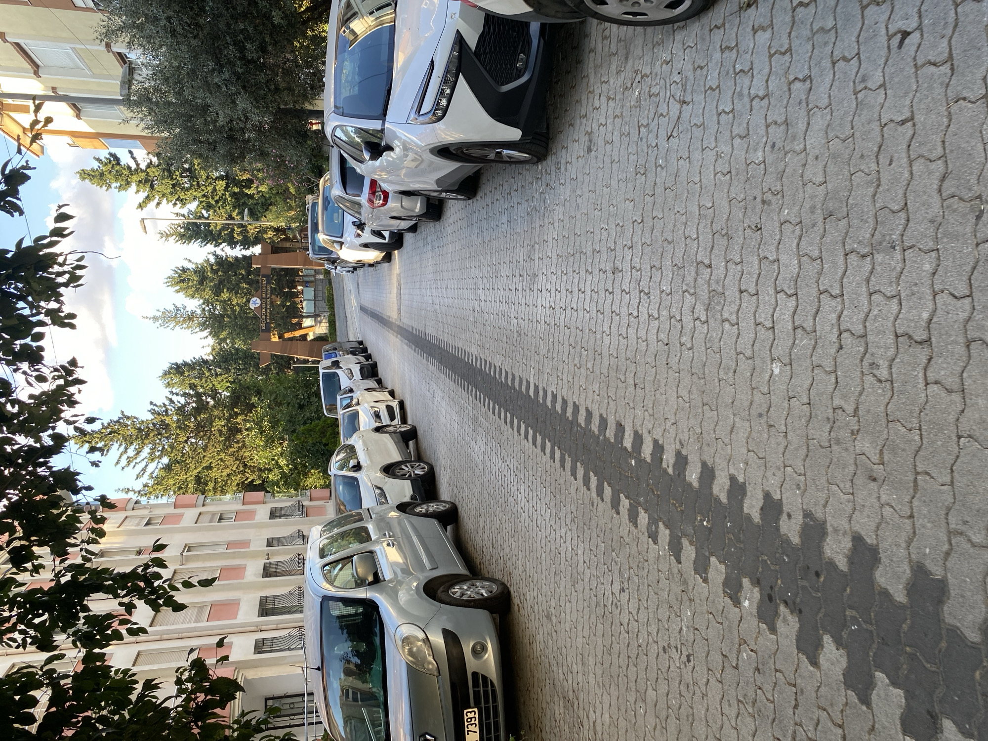 The image depicts a street scene in Küçükçekmece, Turkey, characterized by a row of parked cars on both sides of a paved road. The road surface is made of interlocking grey pavers, with a distinct dark, wet tire track pattern running down the center. On the left side of the road, a silver minivan, identified by the license plate "7393", is parked closest to the foreground, with other vehicles extending further down the street. Behind these cars, a building with pink and cream-colored facade and numerous windows is visible, partially obscured by lush green trees and foliage. On the right side of the road, a line of white cars is parked, including a modern white SUV with a prominent black grille. Further down the street, an archway with signage that appears to read "Küçükçekmece Belediyesi" (Küçükçekmece Municipality) is visible, suggesting this is a public area within the district. Tall, slender evergreen trees are interspersed with the architecture and cars, contributing to a natural urban landscape. The sky above is blue with scattered white clouds, indicating daytime. There are no people visible in the image.