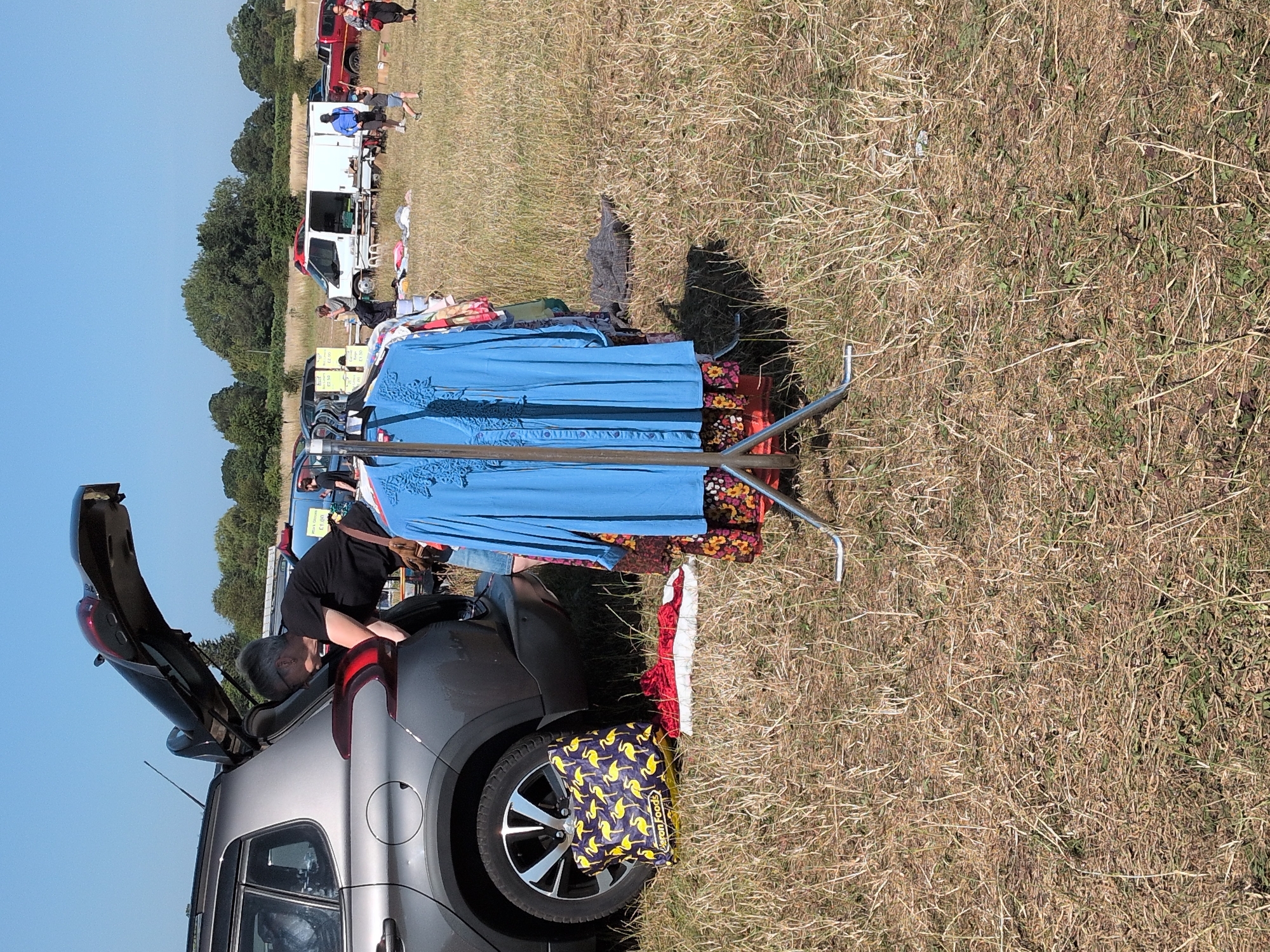 A person is loading items into the trunk of a gray car at an outdoor market. The car's trunk is open, and the person is leaning into it. A rack of clothing hangs in the foreground, and other people and vehicles are visible in the background, suggesting a car boot sale. The ground is covered with dry grass, and the sky is clear and blue. The location is the village of Claverley, United Kingdom.