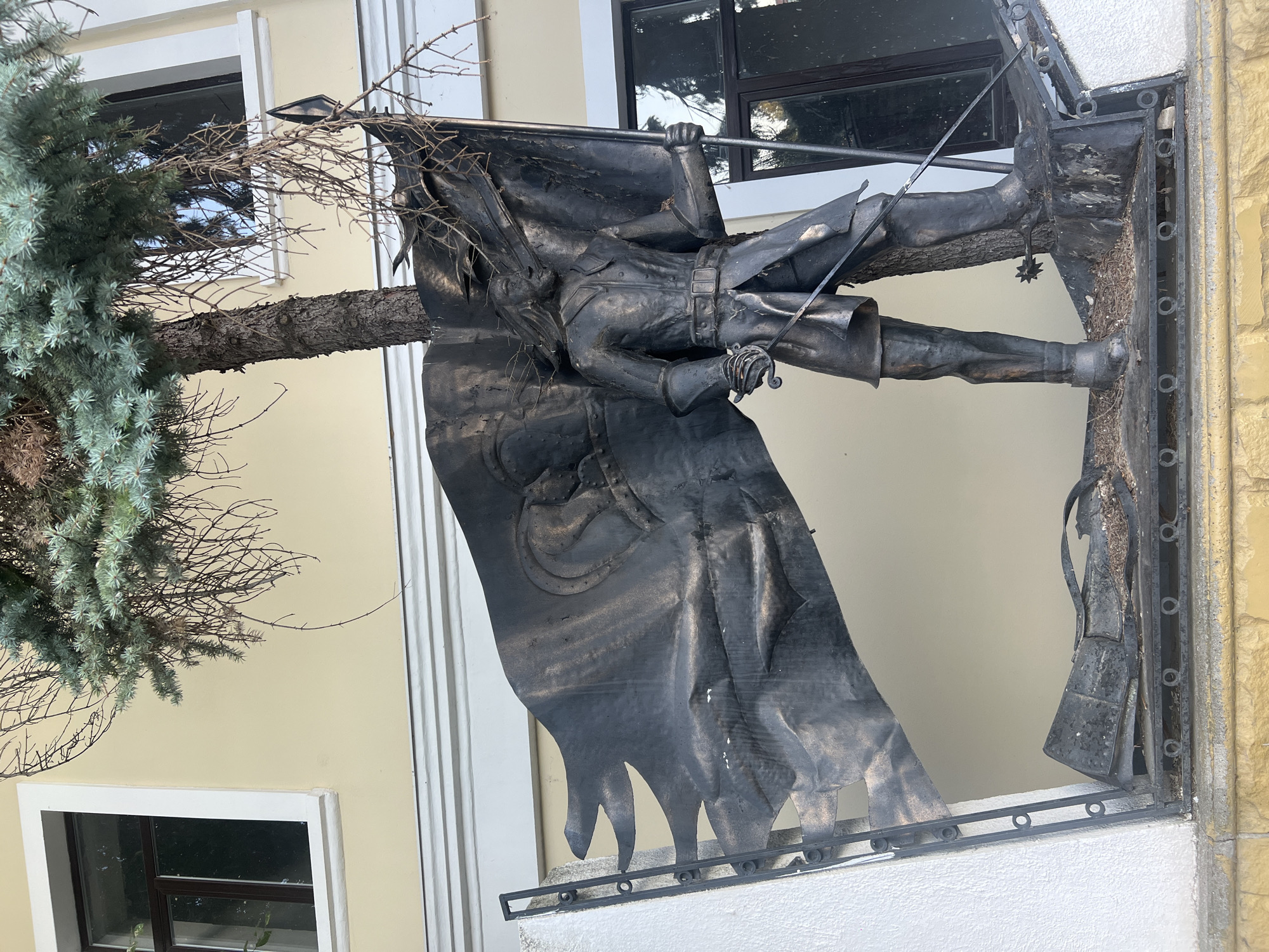 This is a medium-shot, eye-level, outdoor, daytime photograph of a dark metal sculpture depicting a man holding a tattered flag. The sculpture is located in front of a light yellow building with white trim and a window to the left. A tree trunk is integrated into the sculpture, passing through the center of the flag and behind the man.
The man in the sculpture is wearing historical military attire, including a coat, belt, breeches, and boots. He has long hair and holds a sword in his left hand. The flag he is holding is large and appears to be made of metal, with a ripped and wavy design. A crest or emblem is visible on the flag. The base of the sculpture is also made of dark metal, with some debris and dried leaves present. The overall impression of the sculpture is one of movement and possibly conflict, suggested by the tattered flag and the man's stance.
The building in the background has a large window to the right of the sculpture, and another to the far left. The building's facade is a pale yellow color with white architectural details. The foreground shows a stone wall with a textured surface.
The scene is located in the city of Bender, Moldova, possibly at a historical site or memorial.