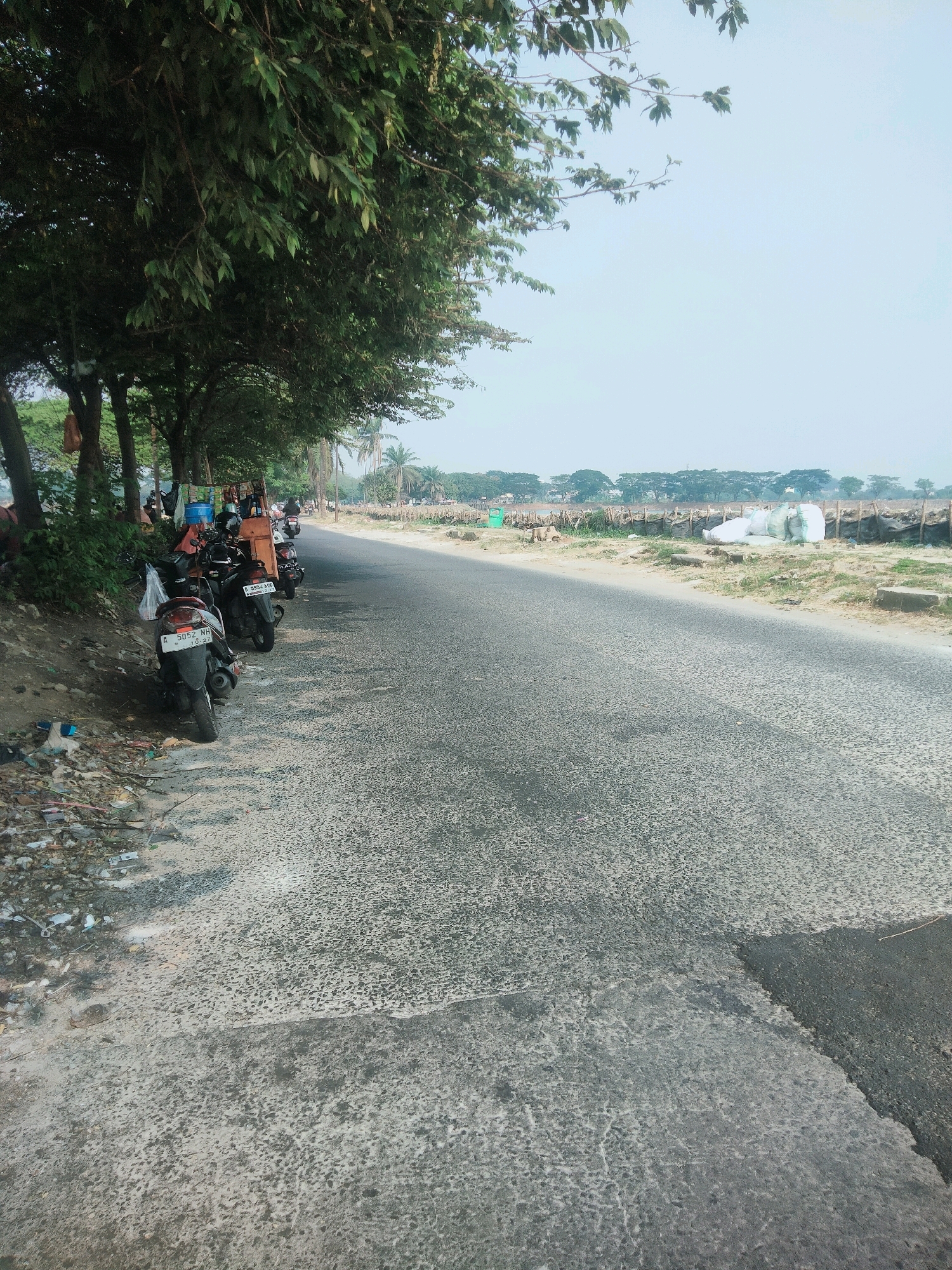 The image depicts a roadside scene in Tangerang, Indonesia. A paved road runs diagonally across the frame, bordered by a dry, unkempt shoulder littered with trash. To the left of the road, a row of parked motorcycles stands under the shade of large trees. Several people are visible near the motorcycles, some appearing to be interacting with a small roadside stall. In the background, across the road, are fields and a line of trees, with some large white sacks visible near a fence. The sky is pale and overcast, suggesting a hazy or possibly polluted atmosphere. The overall impression is one of a utilitarian roadside area, possibly on the outskirts of a more urban environment.