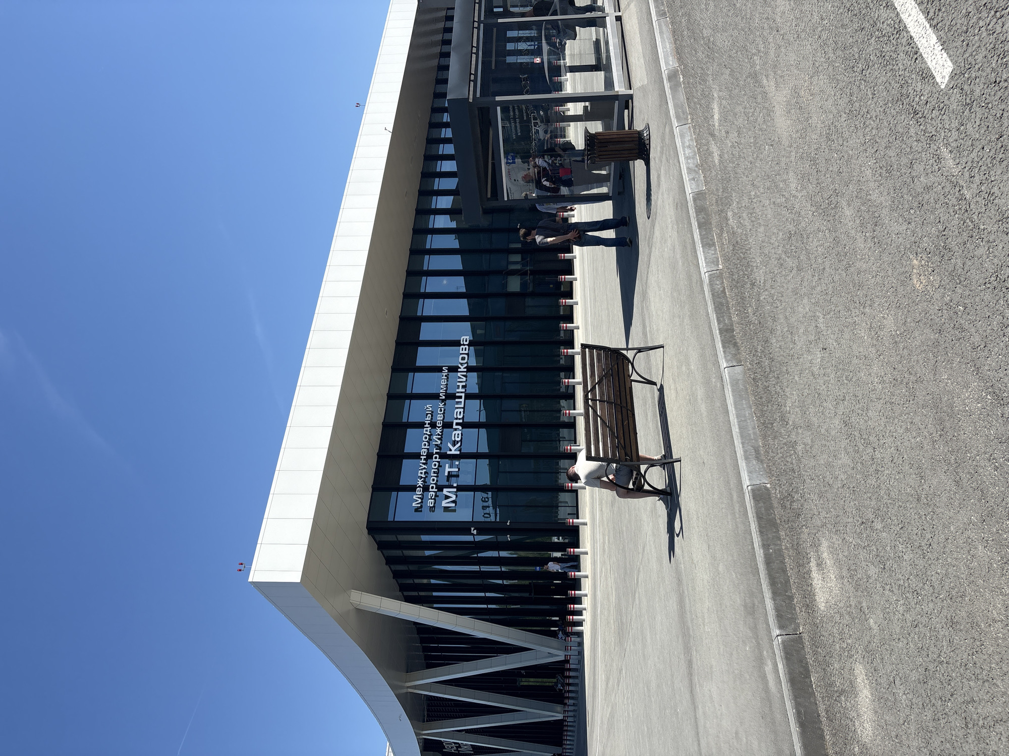 The image shows the exterior of the Izhevsk International Airport named after Mikhail Kalashnikov, located in Novo-Martyanovo, Russia. The building is modern with a white facade and large glass windows bearing the airport's name in Russian. Two people are visible; one stands near the entrance and the other sits on a bench. The surrounding area appears to be paved with concrete and asphalt, including benches and bollards. The sky is clear and blue.